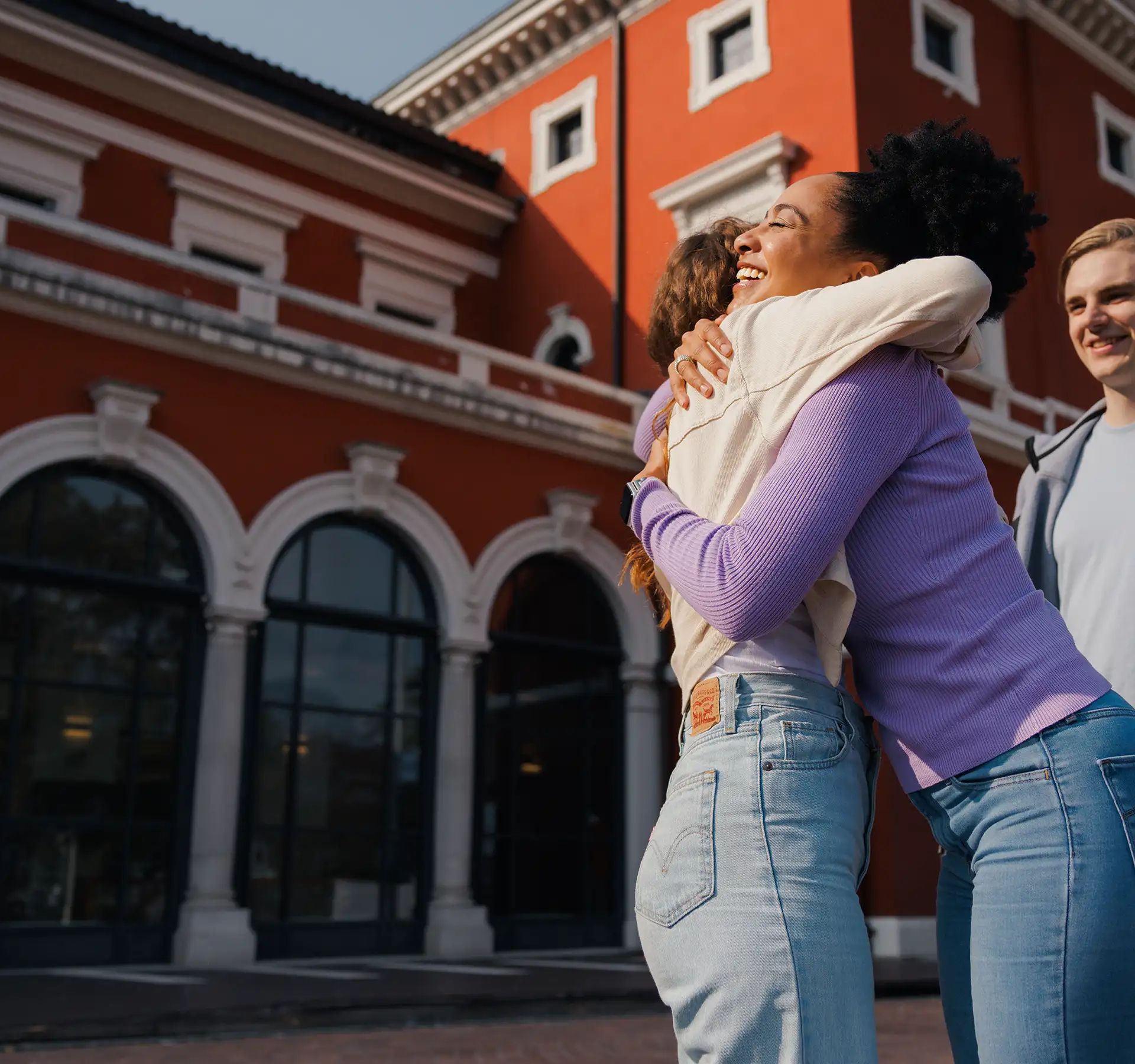 Three young people are standing on a cobblestone square, while the two women embrace happily.