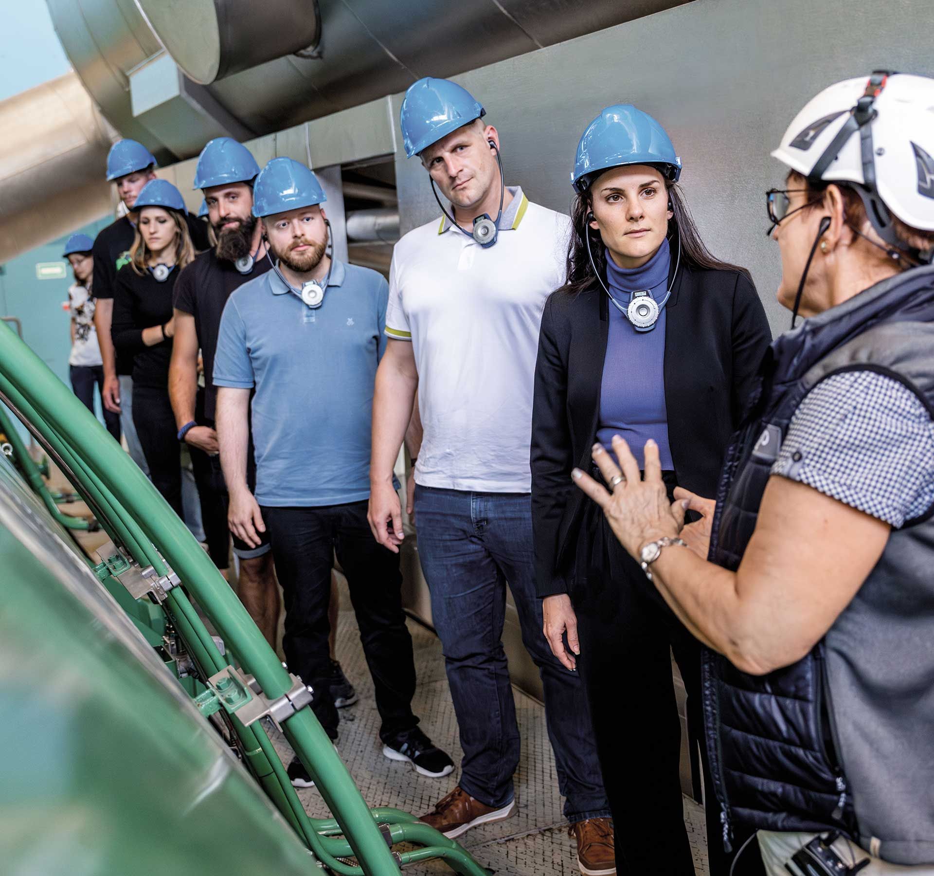 A group of people listen attentively to a guided tour inside the nuclear power plant. Everyone is wearing a helmet and standing next to a green tank. A large silver tube runs above the people.