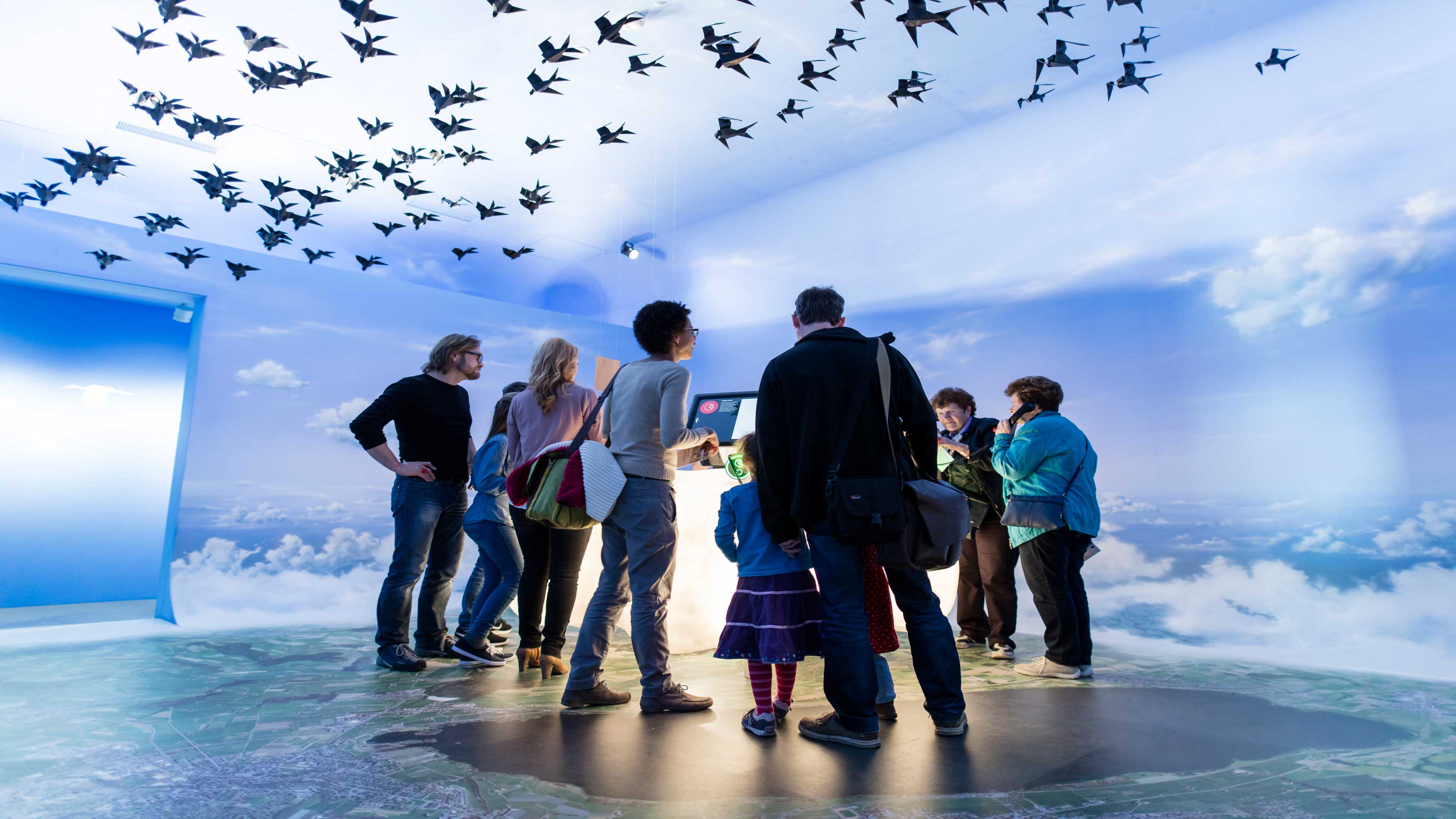 Un groupe se tient dans une salle de la station ornithologique de Sempach. Le sol est une représentation de la perspective d'un oiseau, les murs et le plafond représentent le ciel et des oiseaux en papier sont suspendus au plafond.
