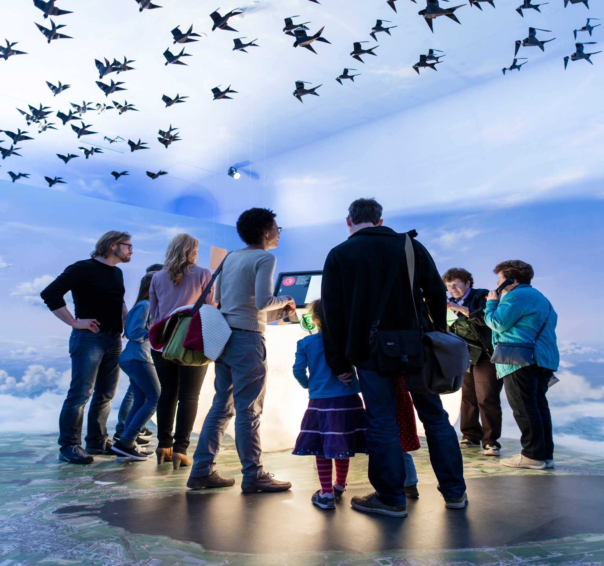 A group is standing in a room at the Sempach ornithological station. The floor is a picture from a bird's eye view, the walls and ceiling show the sky and paper birds hang from the ceiling.
