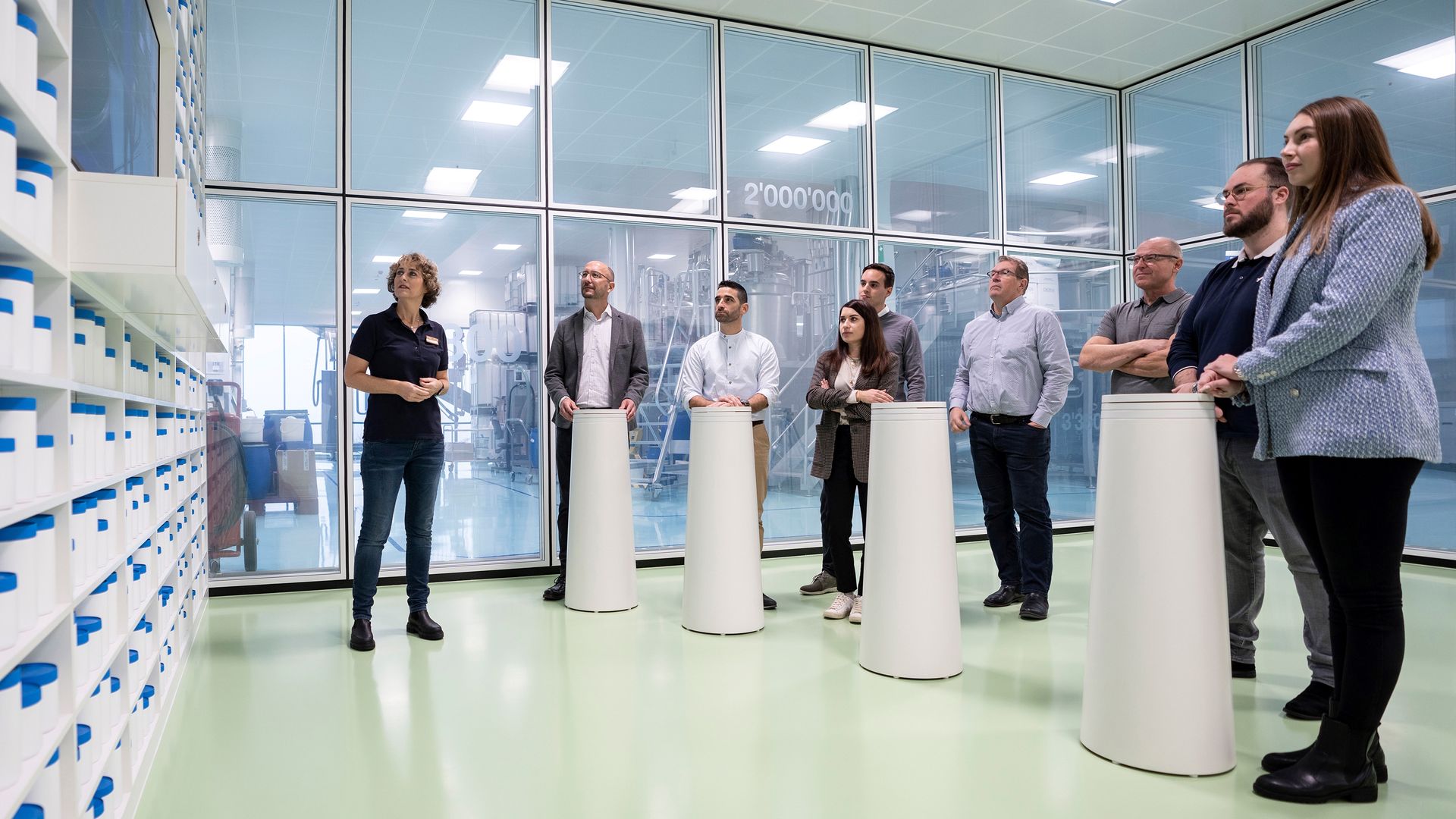 Un gruppo si trova in una stanza con pareti di vetro. Su una parete, fino al soffitto, c'è uno scaffale con contenitori rotondi di diverse dimensioni, bianchi e con il coperchio blu. Il gruppo guarda un film su uno schermo a parete.