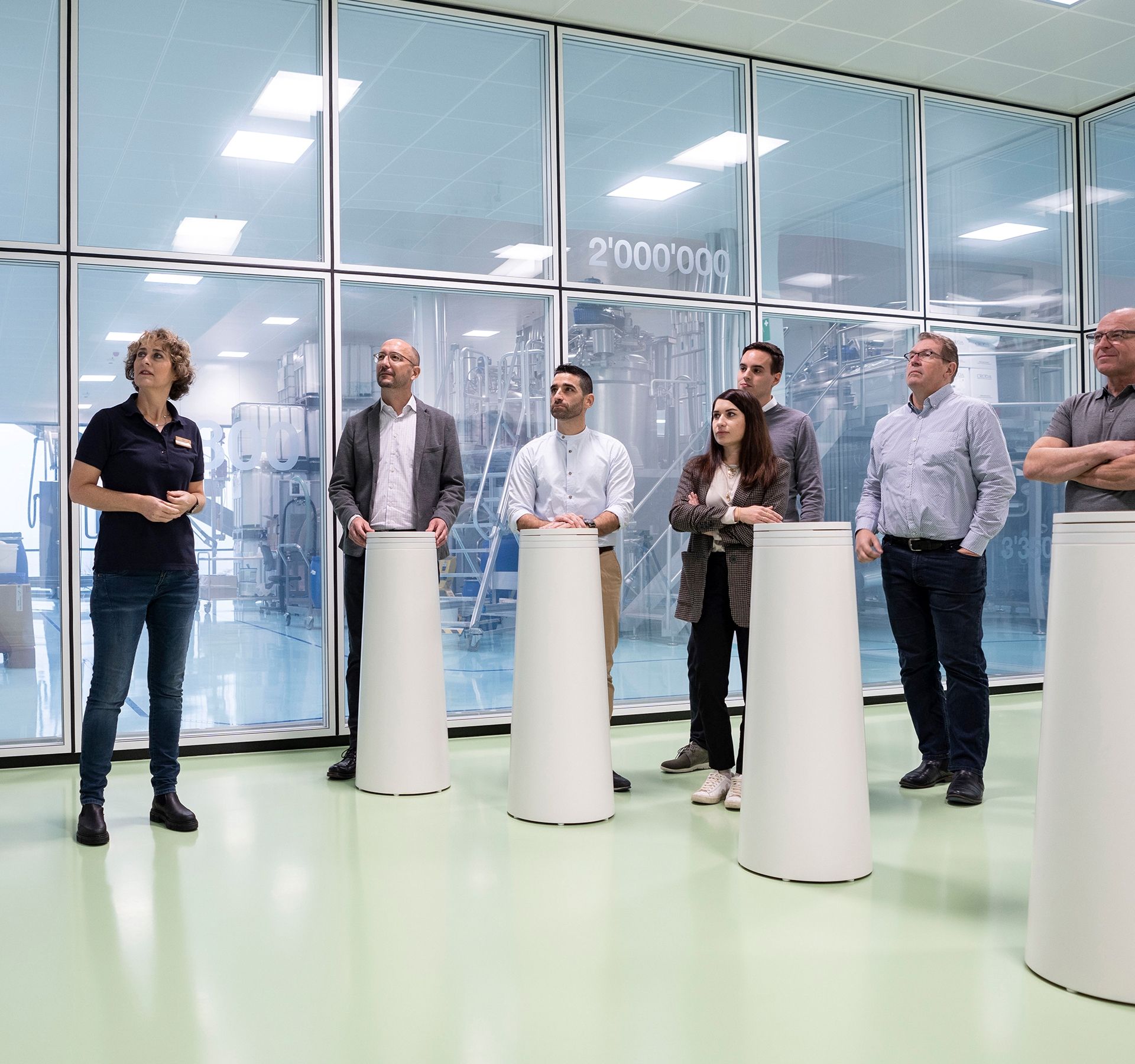 A group stands in a room with glass walls. On one wall up to the ceiling is a shelf with different sized round, white containers with blue lids. The group watches a film on a screen on the wall described above.