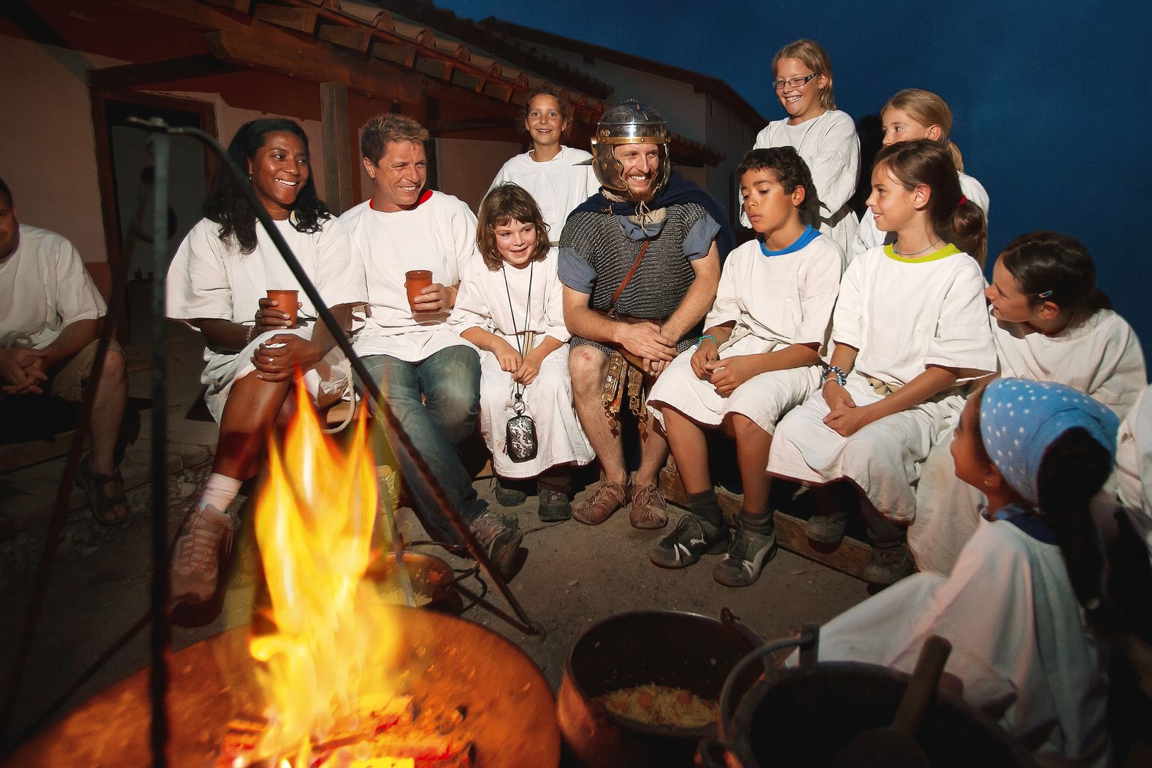 A group is sitting around a fire. A large part of the group is dressed in white tops. One man is dressed as a legionnaire. 