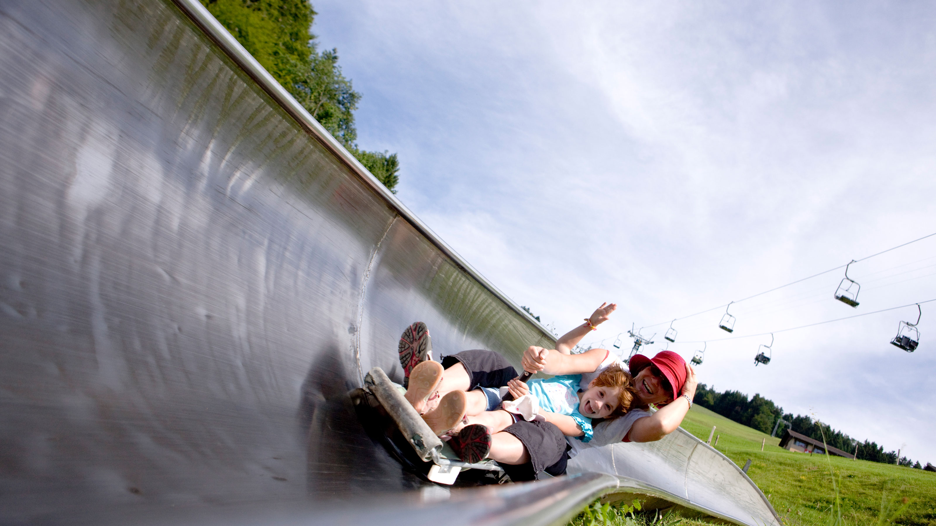 A laughing child and a woman ride down a summer toboggan run together, while a chairlift floats across a green meadow in the background.