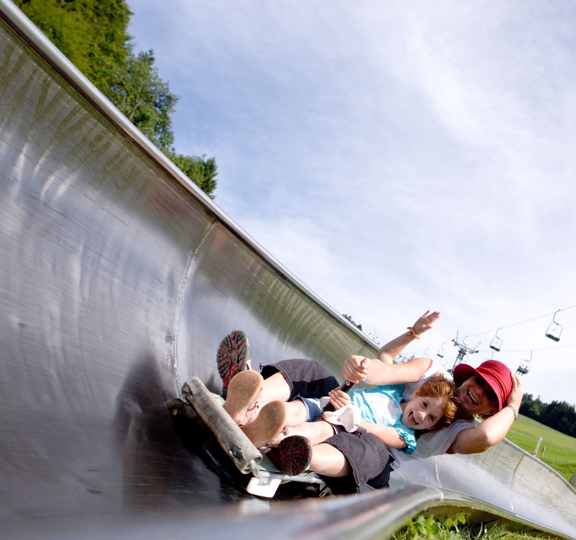 A laughing child and a woman ride down a summer toboggan run together, while a chairlift floats across a green meadow in the background.
