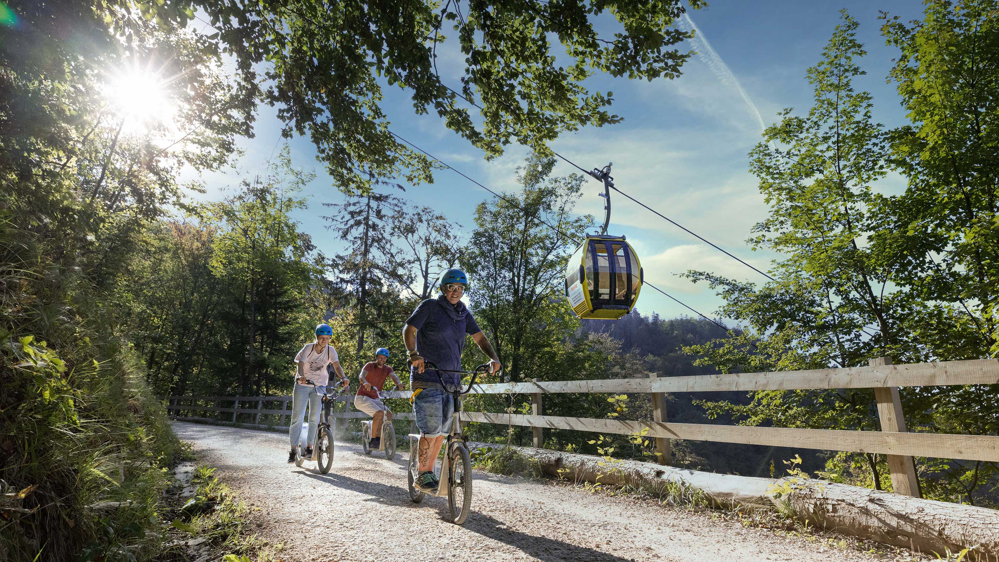 Drei Personen flitzen mit den Trottis die Strasse hinunter. Die Sonne scheint durch die Blätter des Waldes auf der linken Seite und rechts im Hintergrund sieht man die Gondel für auf den Berg.