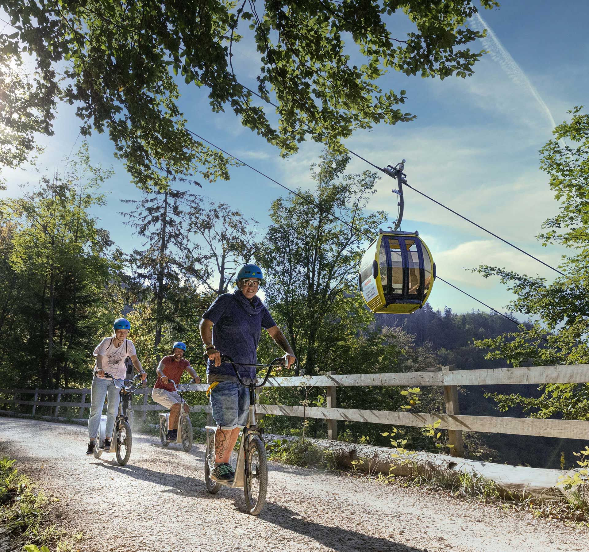 Tre persone sfrecciavano sulla strada in scooter. Il sole illumina le foglie del bosco sulla sinistra e sullo sfondo, a destra, si vede la funivia che sale sulla montagna.