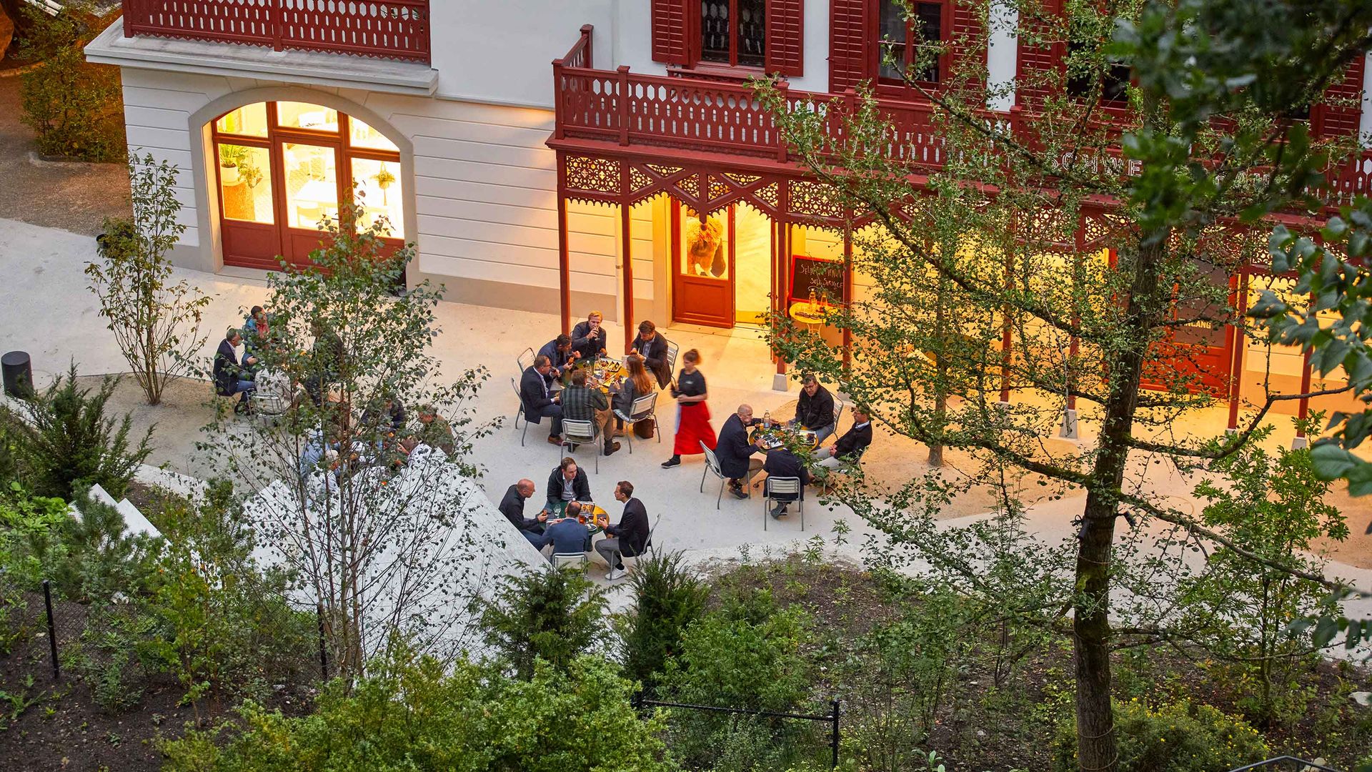 Eine bunt gemischte Gruppe von Menschen sitzt vor einem Gebäude zusammen an Tischen und geniesst ein Apéro.