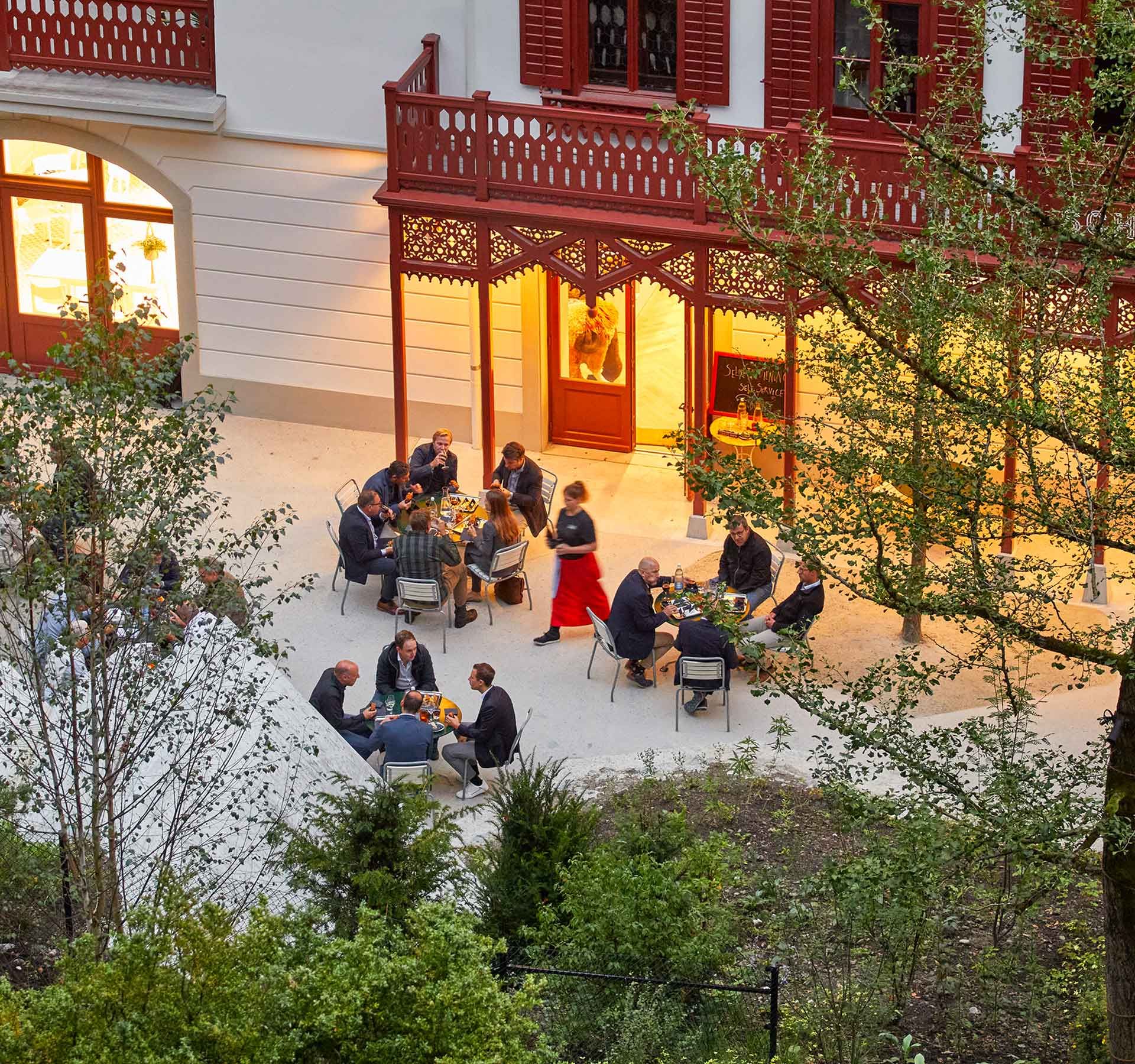 Eine bunt gemischte Gruppe von Menschen sitzt vor einem Gebäude zusammen an Tischen und geniesst ein Apéro.