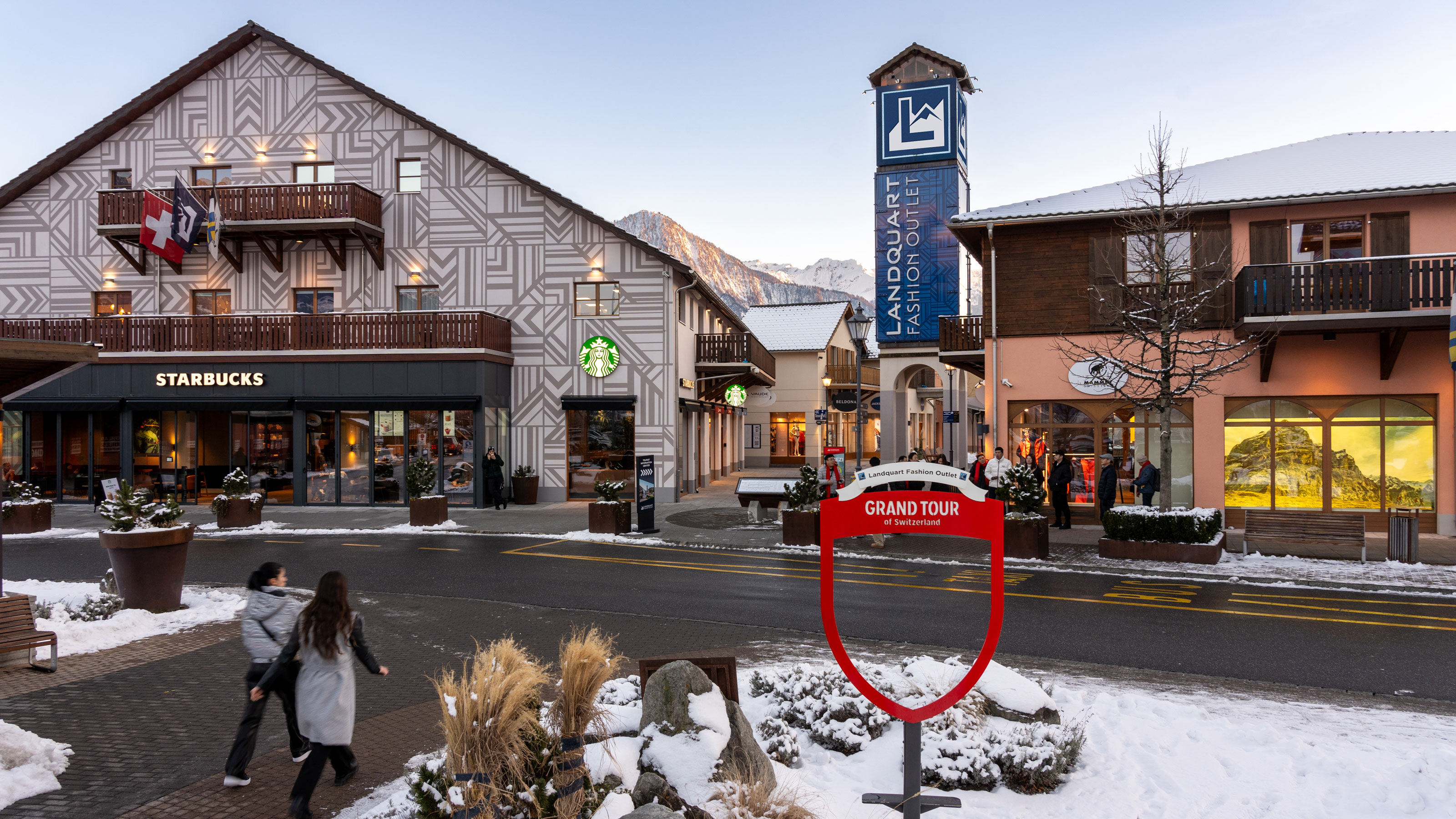 L’immagine mostra il Landquart Fashion Outlet innevato in Svizzera con uno Starbucks e il cartello della “Grand Tour of Switzerland”
