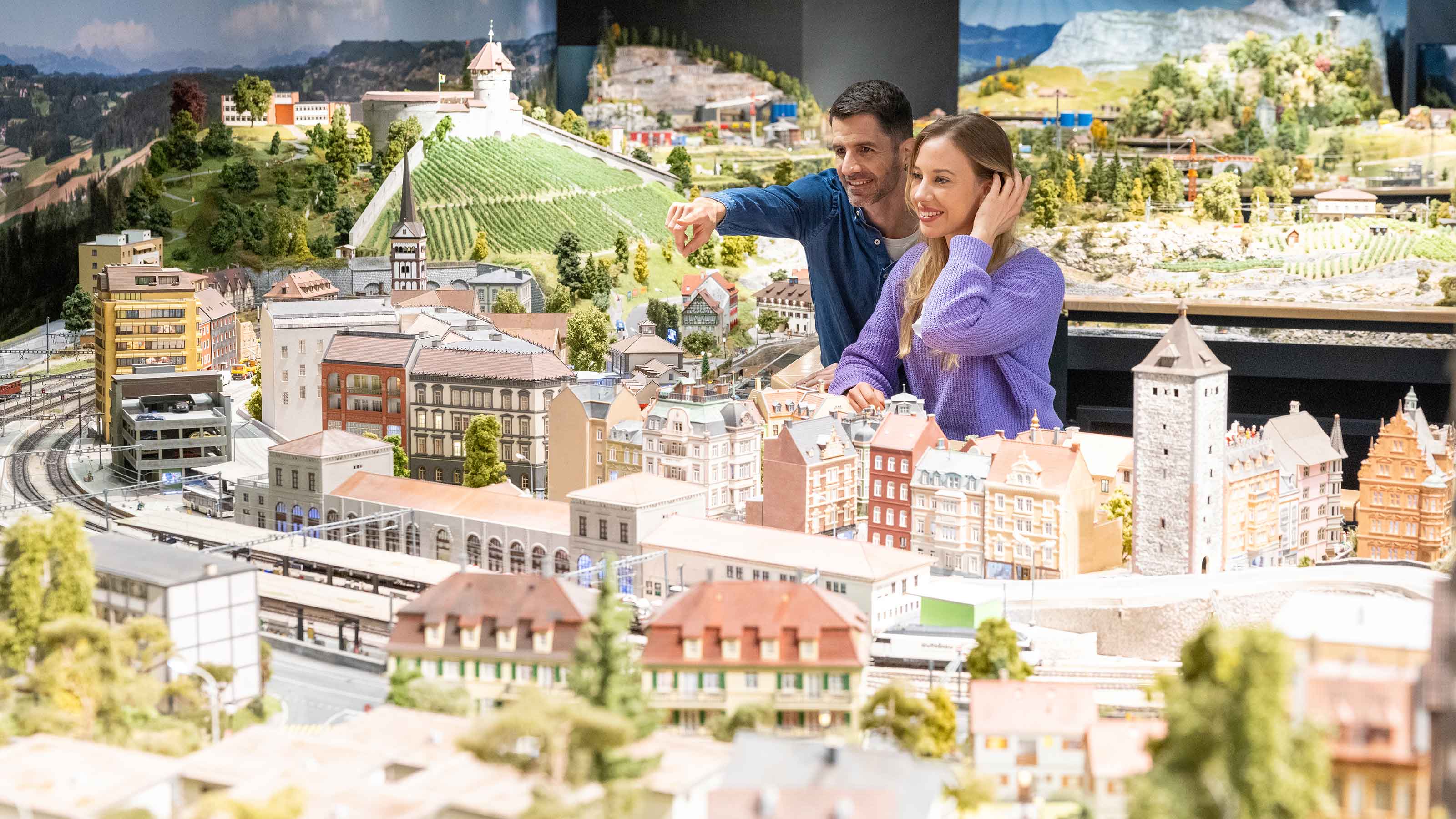 A man and a woman look at the miniature world. The man stands slightly behind the woman and points at something with his finger.