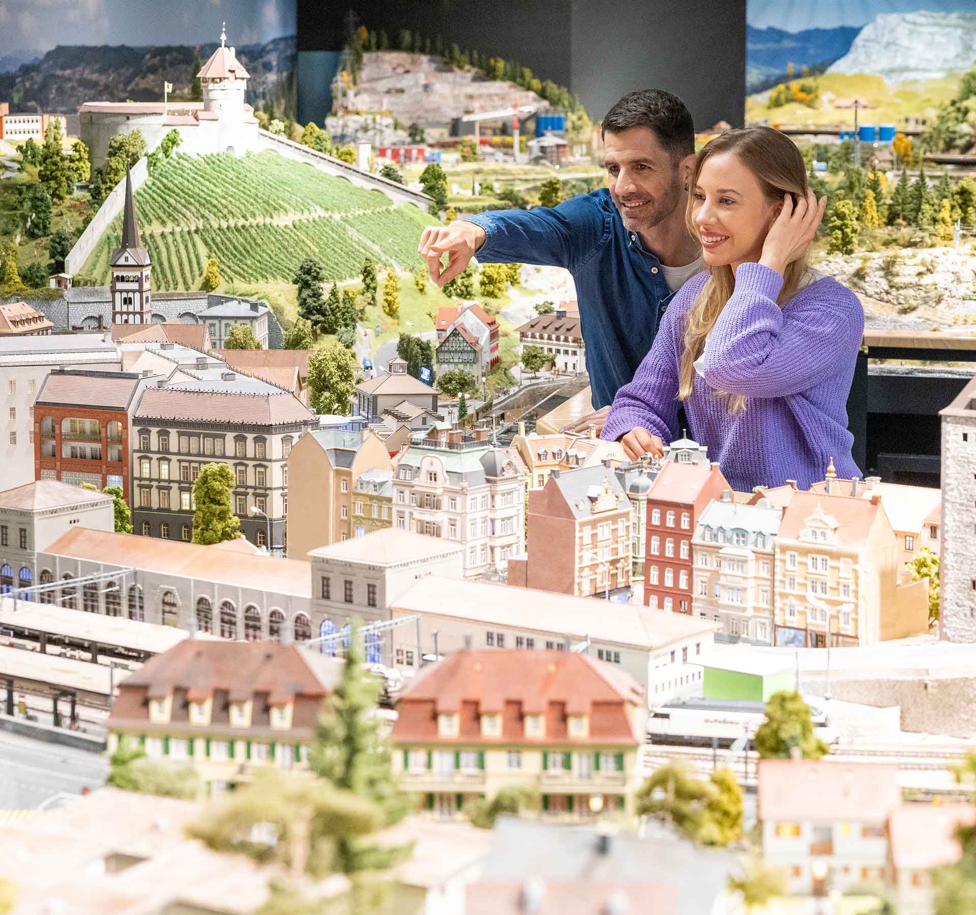 Un homme et une femme regardent le monde miniature. L'homme se tient un peu derrière la femme et montre quelque chose du doigt.