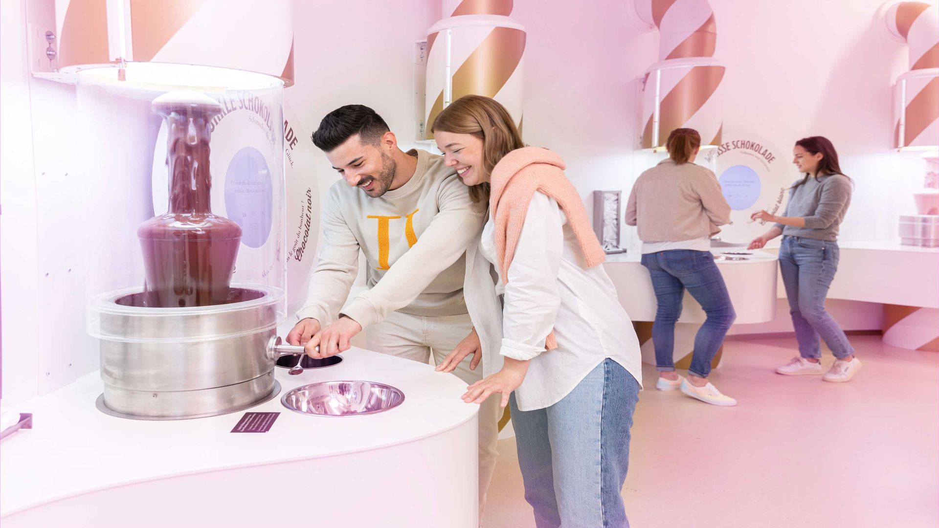 A woman and a man taste chocolate from a small chocolate fountain. In the background, other people are tasting chocolate.