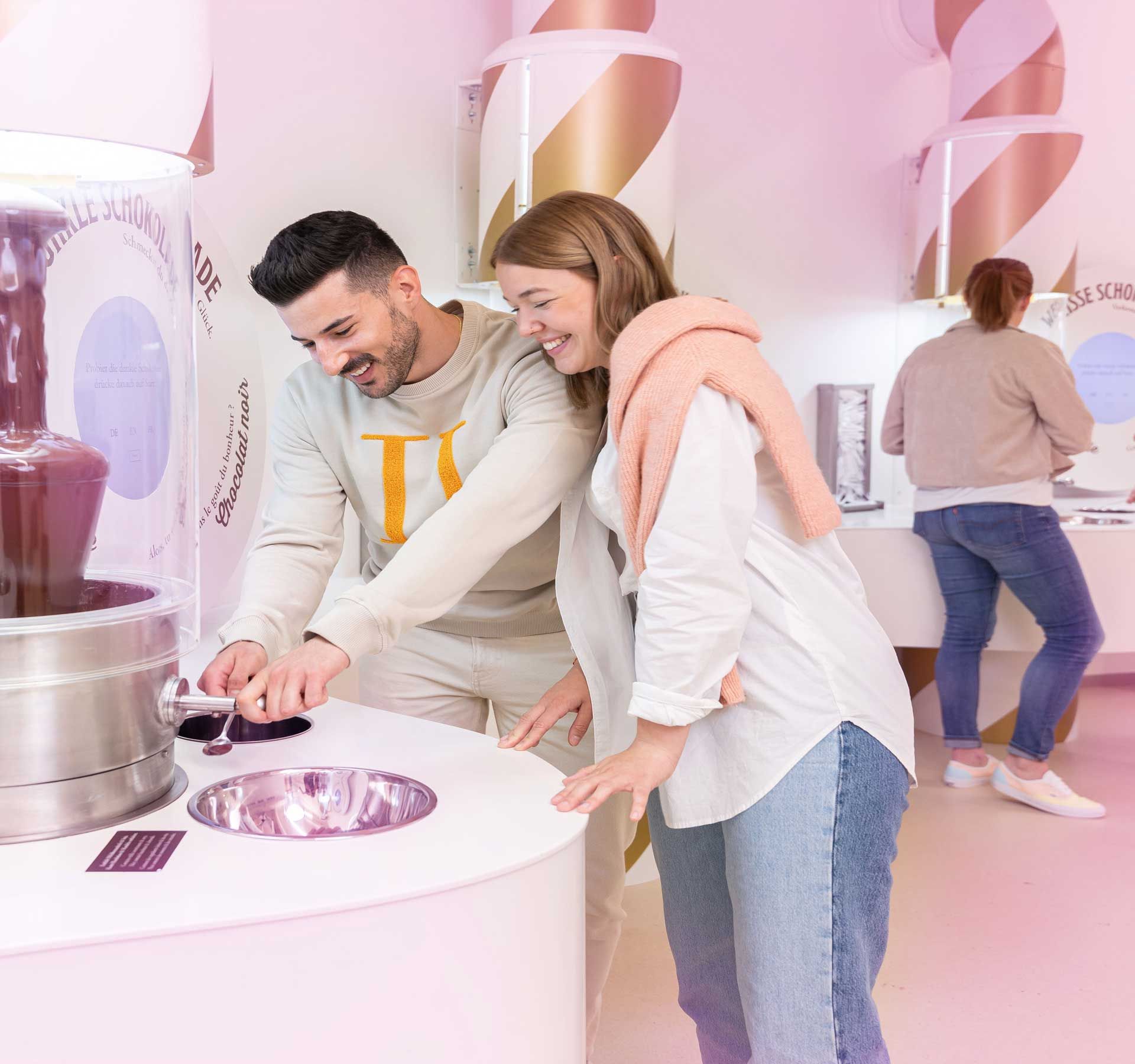Une femme et un homme goûtent du chocolat à une petite fontaine à chocolat. En arrière-plan, d'autres personnes dégustent du chocolat.
