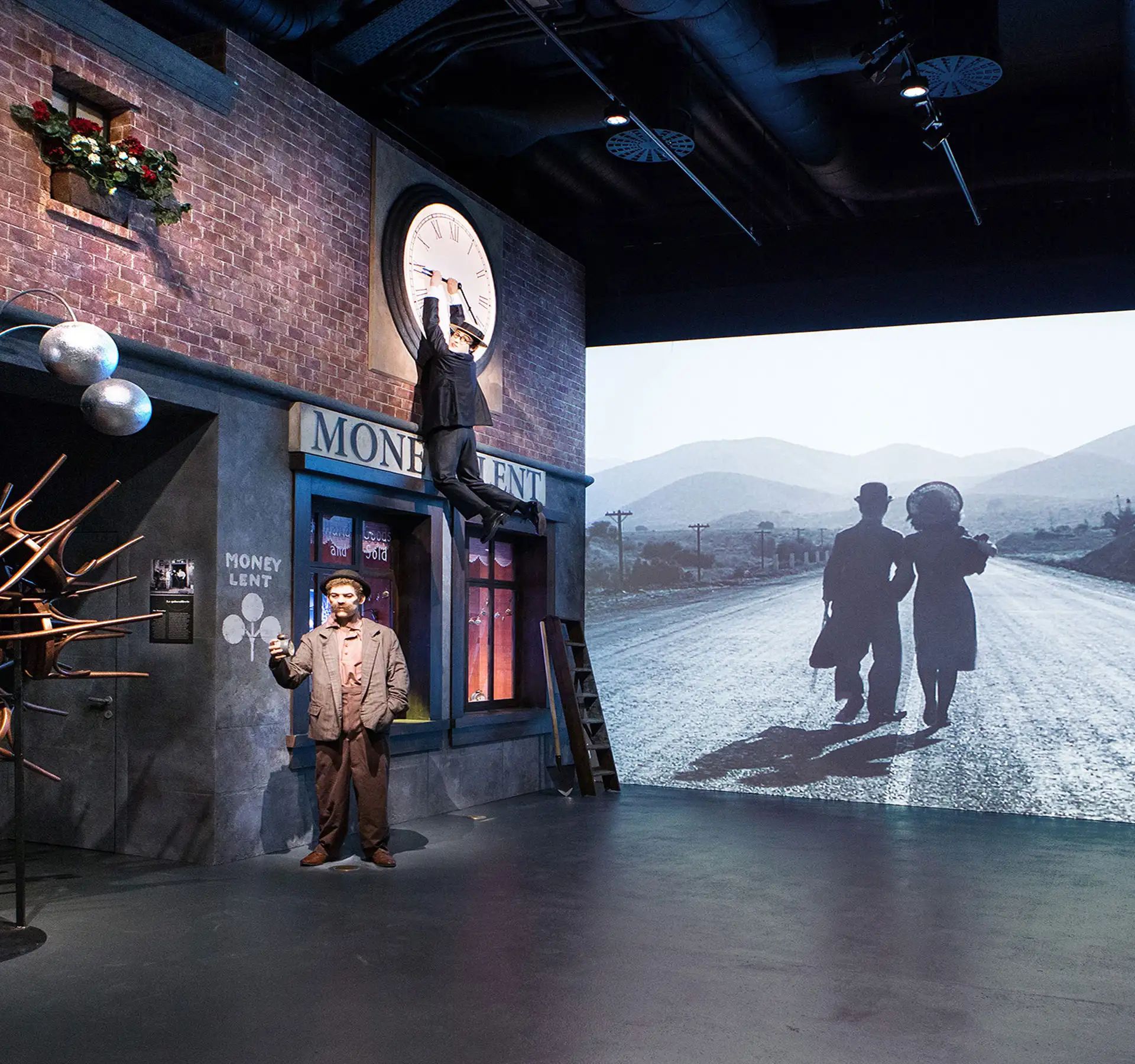 In una mostra museale si possono vedere un edificio in mattoni, un pupazzo vestito da Charlie Chaplin in una strada e un filmato in bianco e nero in cui due persone camminano verso un orizzonte montuoso.