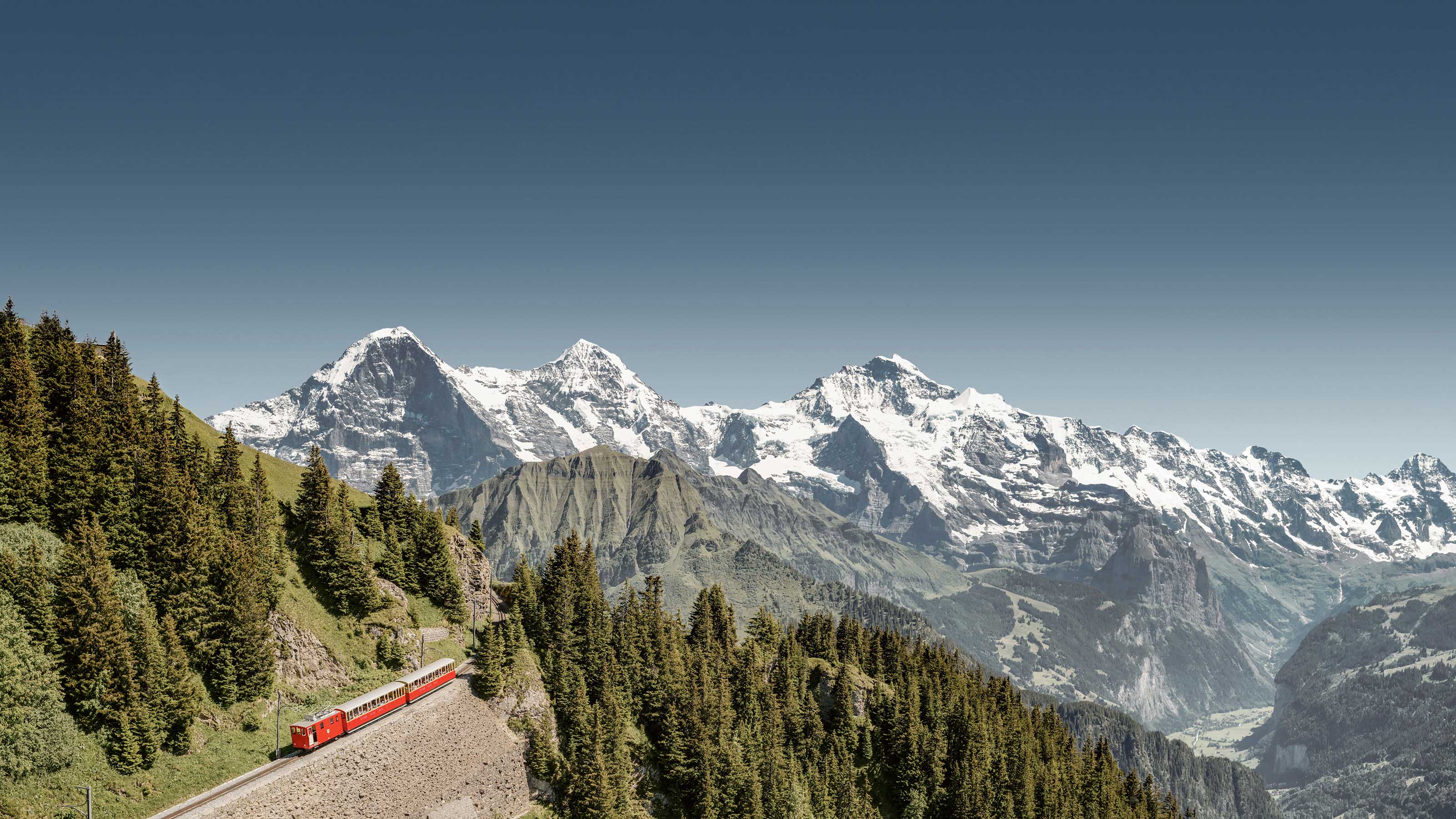 Luftaufnahme der Zahnrad welche den Berg hinochfährt. Im Hintergrund sind verschneite Berggipfel Eiger, Mönch und Jungfrau zu sehen.