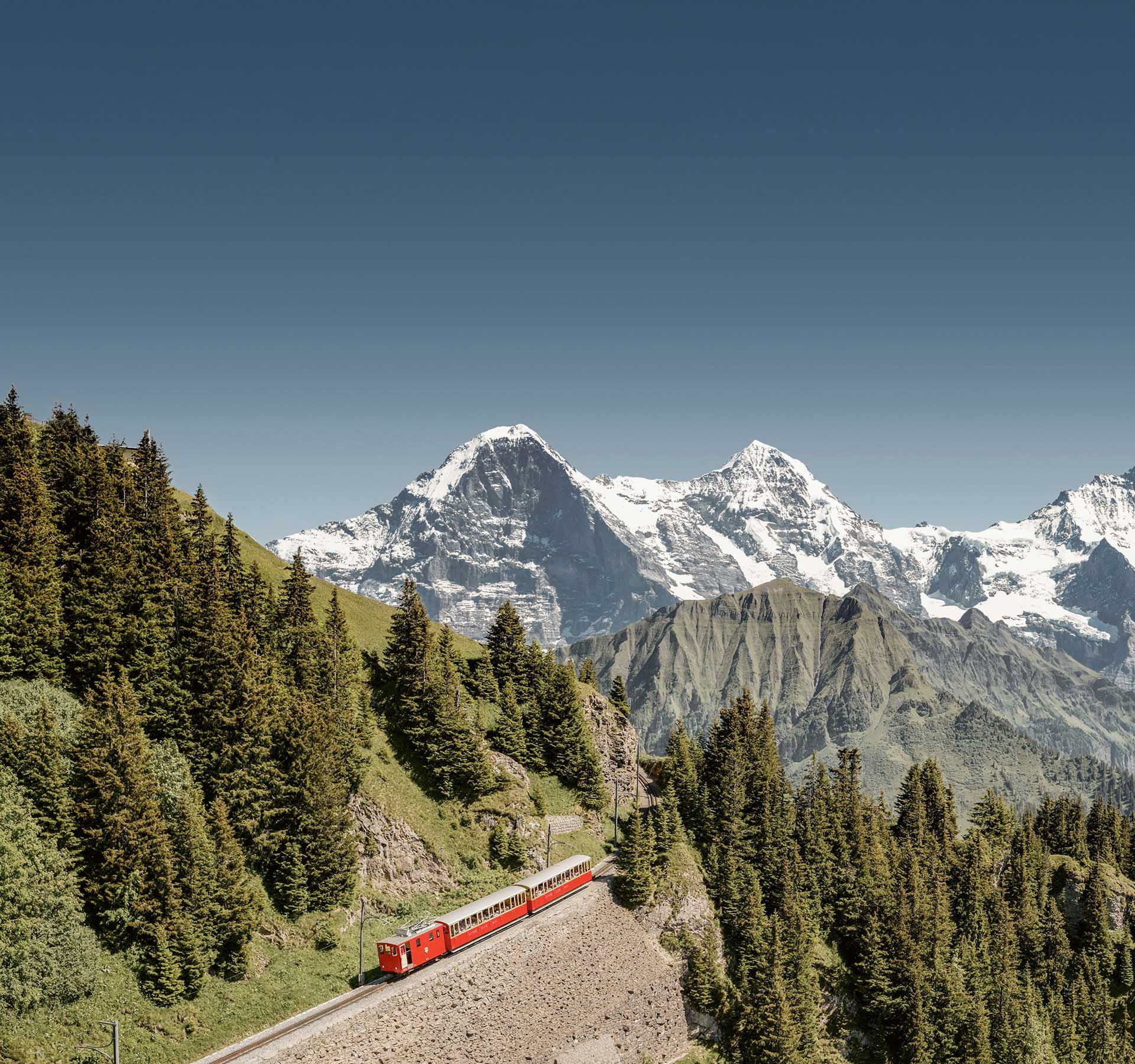 Luftaufnahme der Zahnrad welche den Berg hinochfährt. Im Hintergrund sind verschneite Berggipfel Eiger, Mönch und Jungfrau zu sehen.