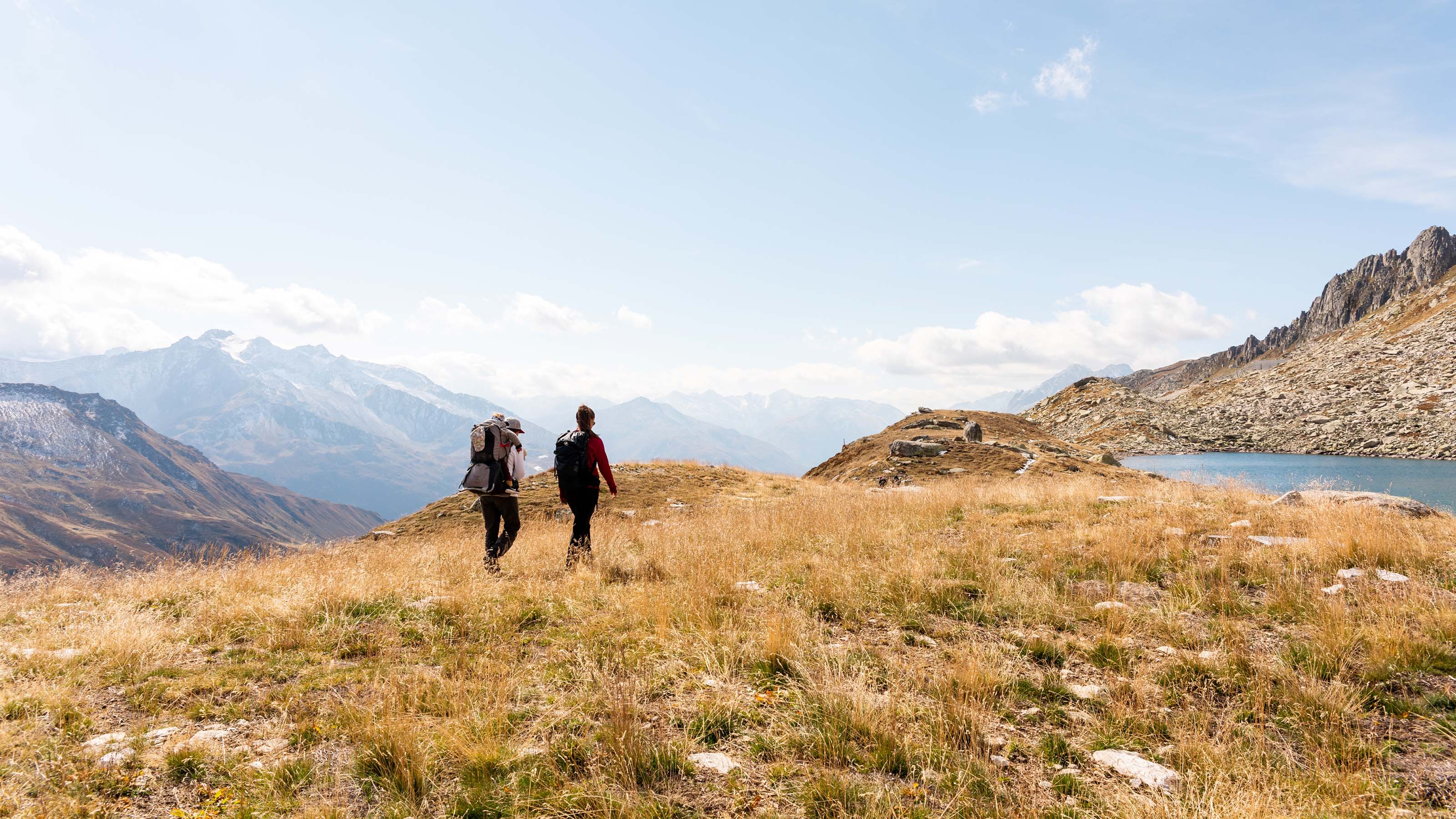 Due escursionisti attraversano un prato vicino a un piccolo lago di montagna nella regione di Andermatt. È una bella giornata estiva di sole e la vista spazia fino alle cime più lontane.