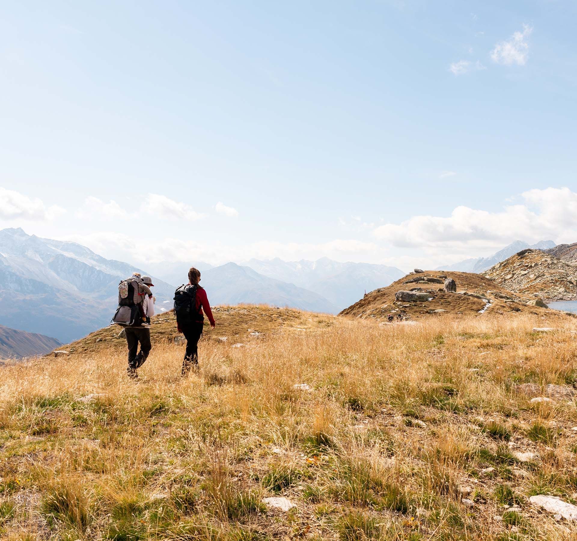 Two hikers cross a meadow near a small mountain lake in the Andermatt region. It is a beautiful, sunny summer day and the view reaches far to distant peaks.