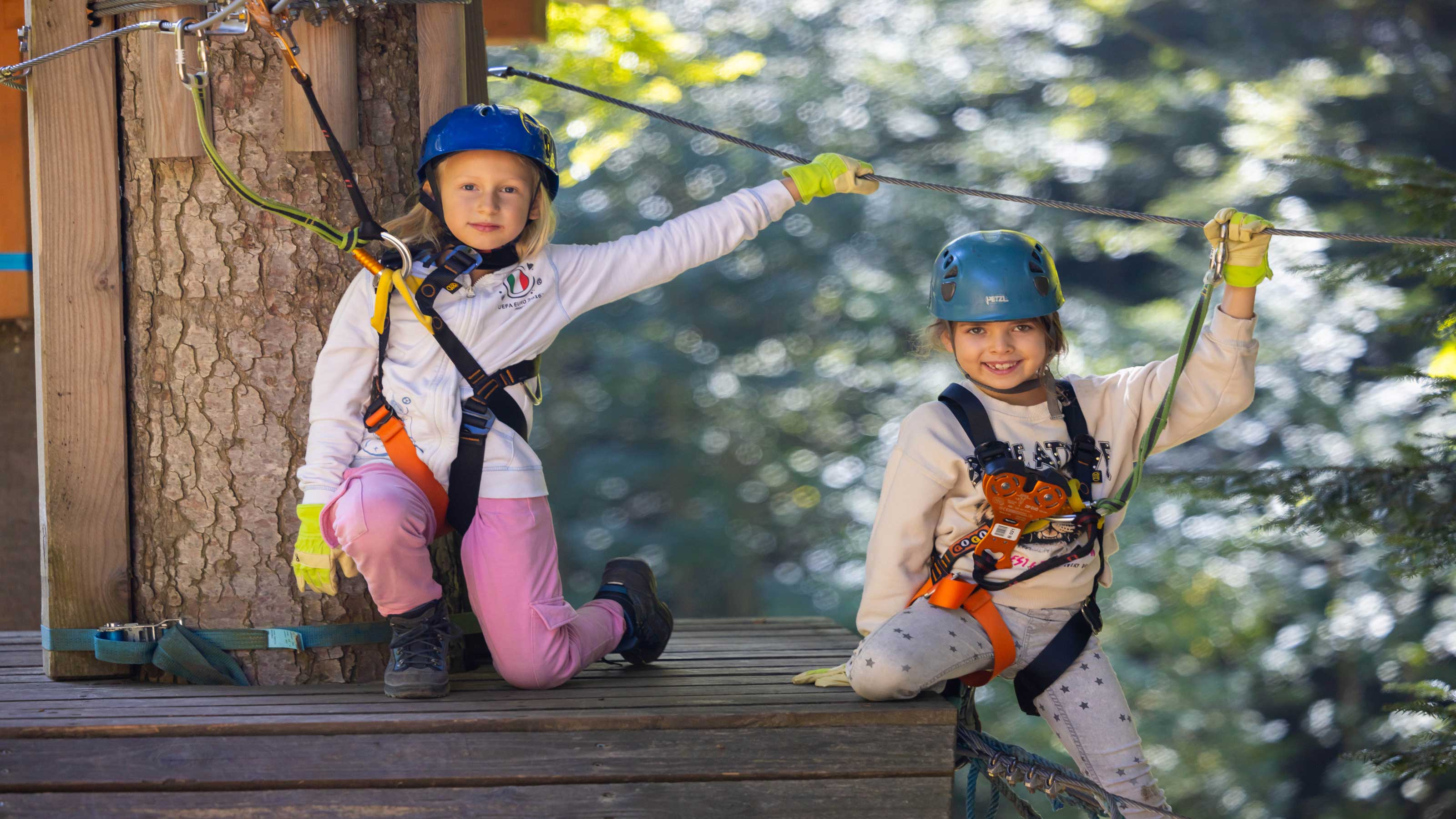 Deux enfants dans un parc d'aventure en forêt portent des casques et des ceintures de sécurité, souriant joyeusement à la caméra.