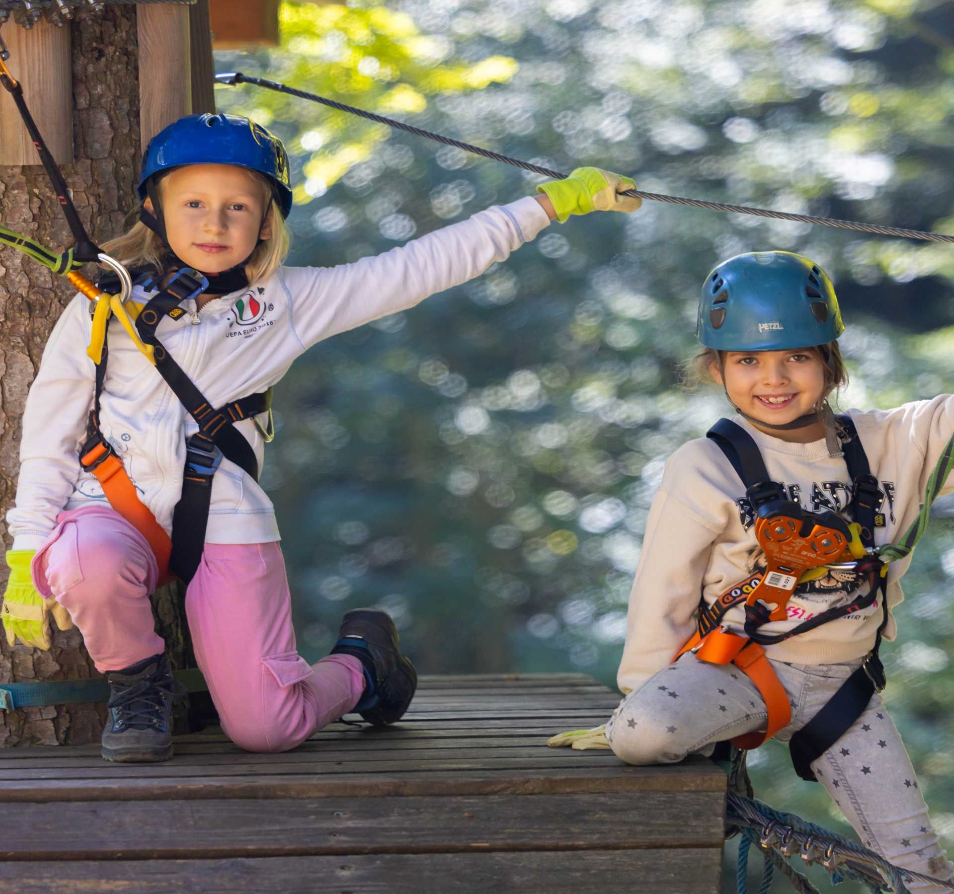 Two children in the forest ropes course wearing helmets and safety harnesses, smiling happily at the camera.