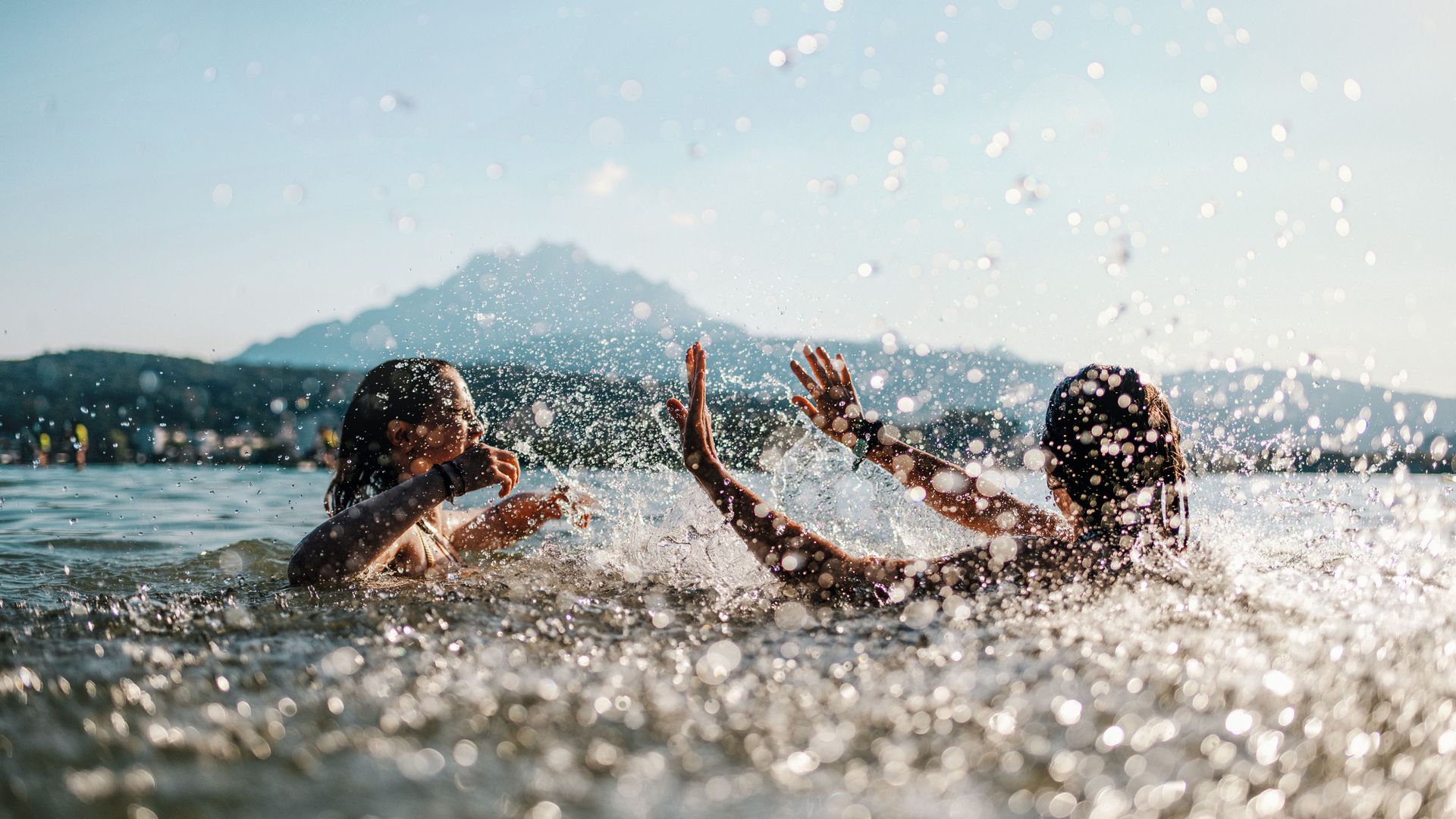Due ragazze si godono le temperature estive e fanno il bagno nel lago dei Quattro Cantoni. Si schizzano a vicenda con l'acqua. I raggi del sole si infrangono sulle gocce d'acqua.