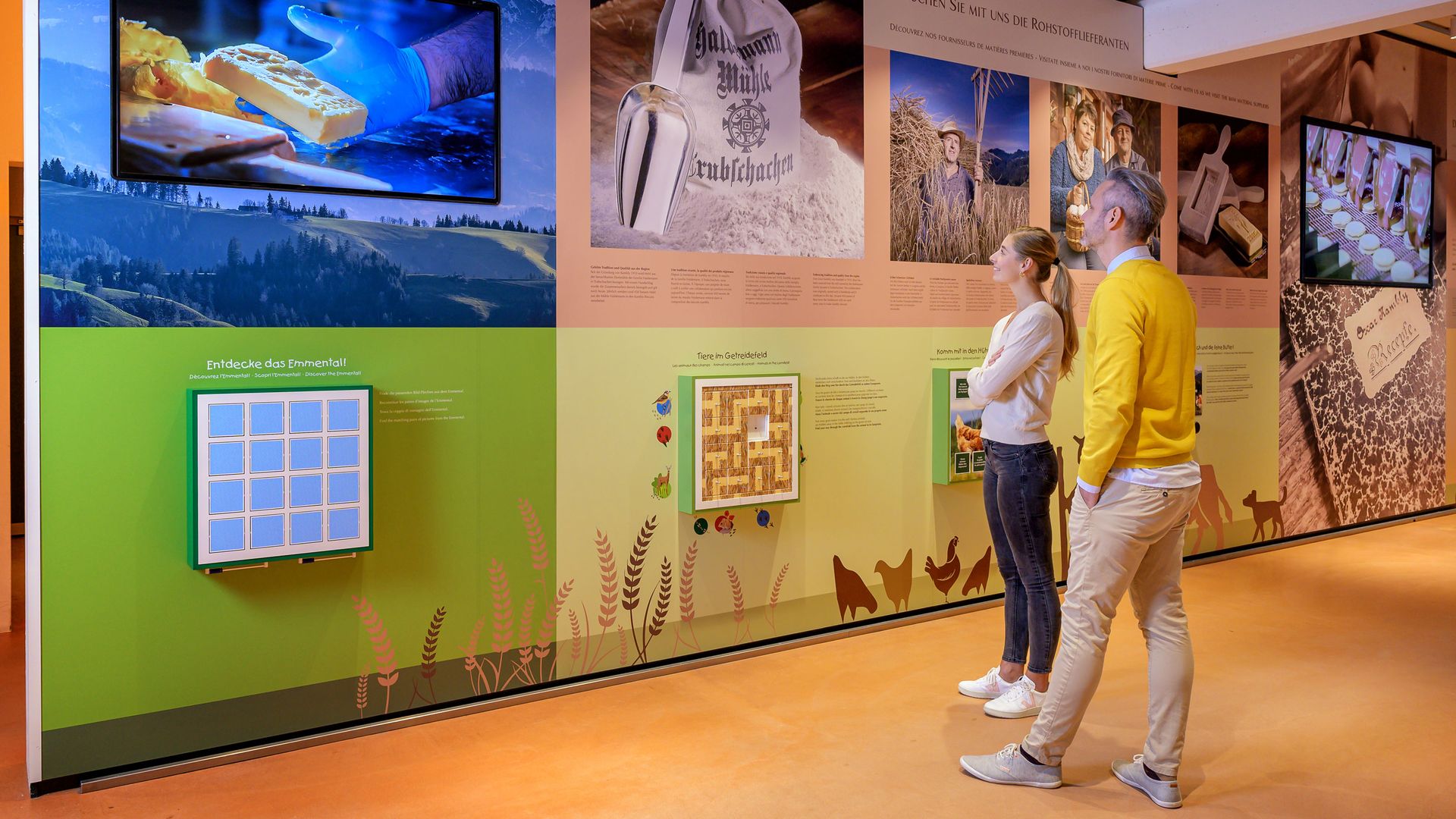 Two people looking at an exhibition with texts, images, and interactive displays about the origin of Kambly products in a museum or visitor center.