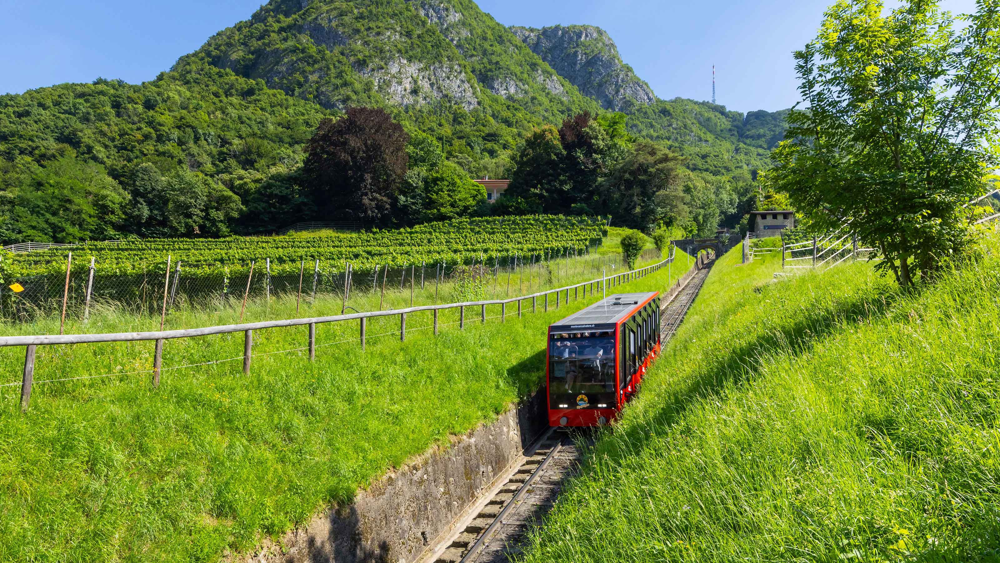 Una funicolare rossa che parte dal Monte San Salvatore attraversa un paesaggio verde con vigneti e montagne sullo sfondo.
