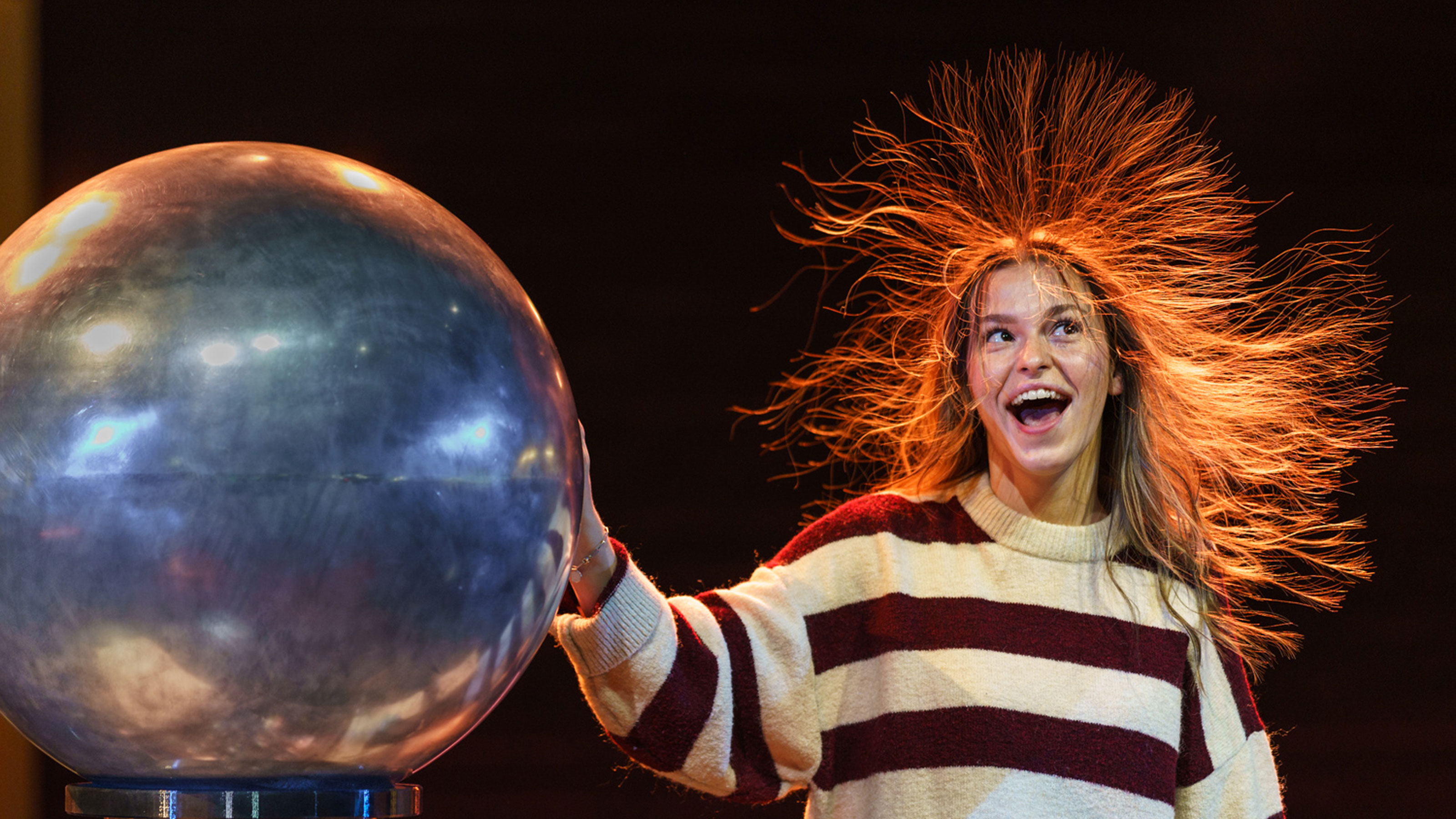 Person with long hair and striped sweater standing against dark background touching a large metal sphere causing hair to stand up due to static electricity