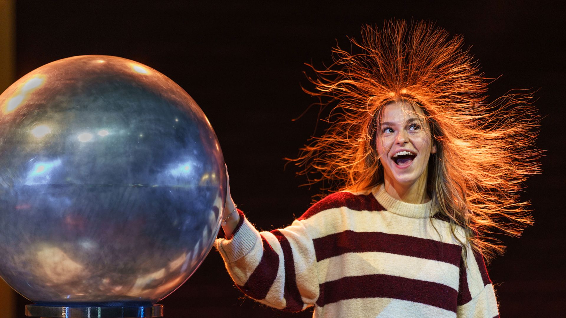 Person with long hair and striped sweater standing against dark background touching a large metal sphere causing hair to stand up due to static electricity