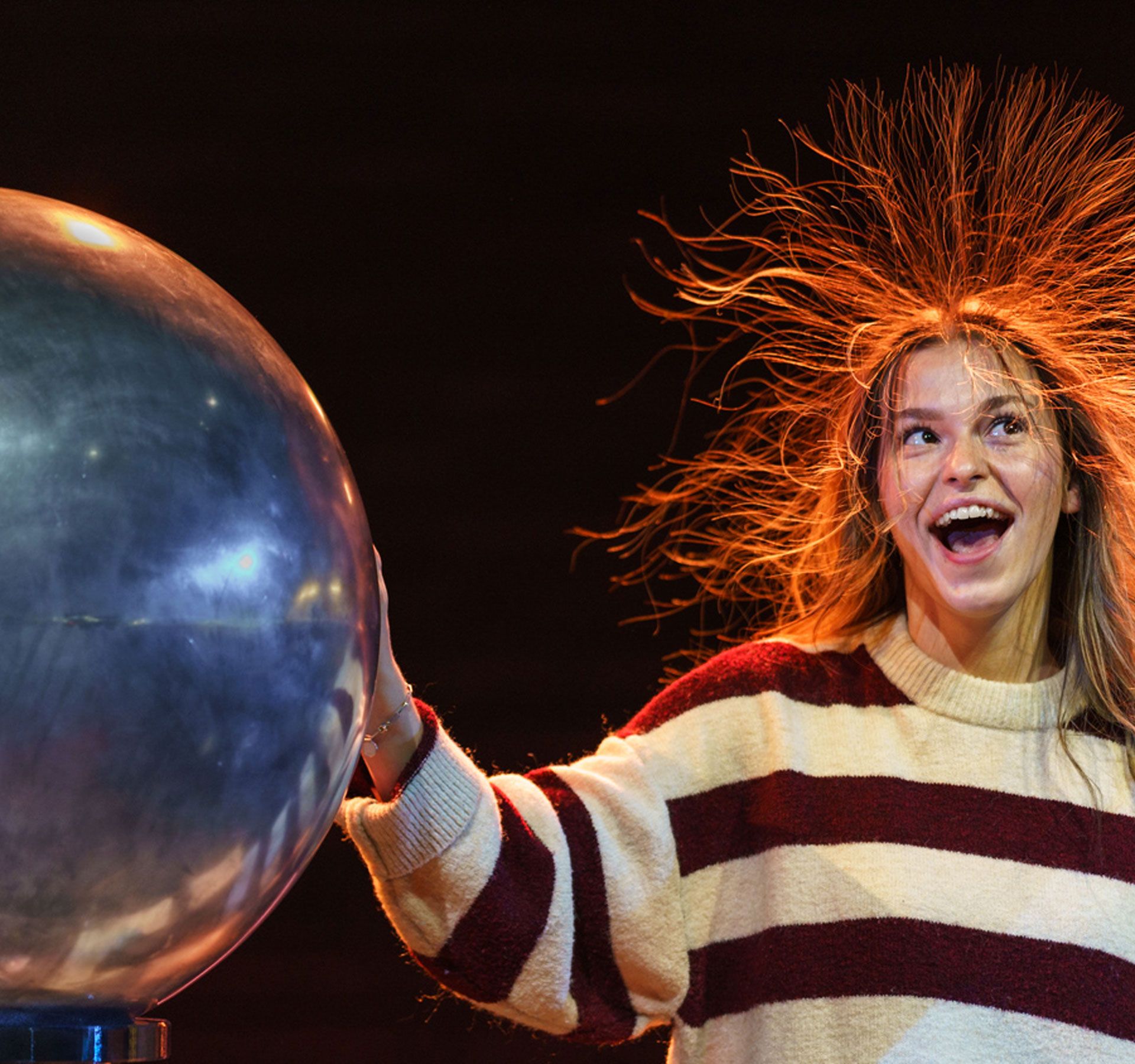 Person with long hair and striped sweater standing against dark background touching a large metal sphere causing hair to stand up due to static electricity