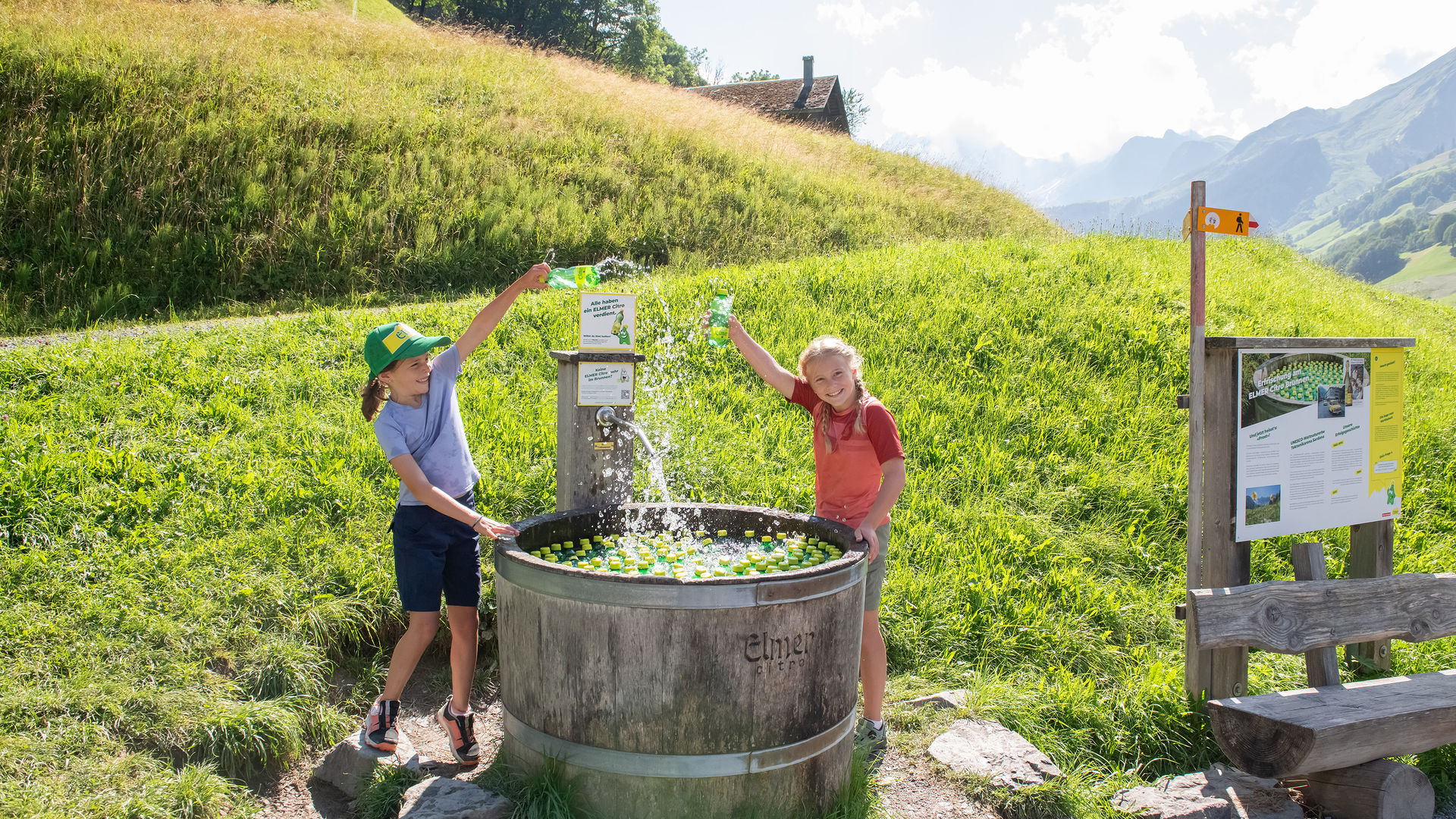 Deux jeunes filles tirent joyeusement une petite bouteille d'Elmer Citro d'une grande fontaine dans laquelle flottent de nombreuses autres bouteilles d'Elmer Citro...
