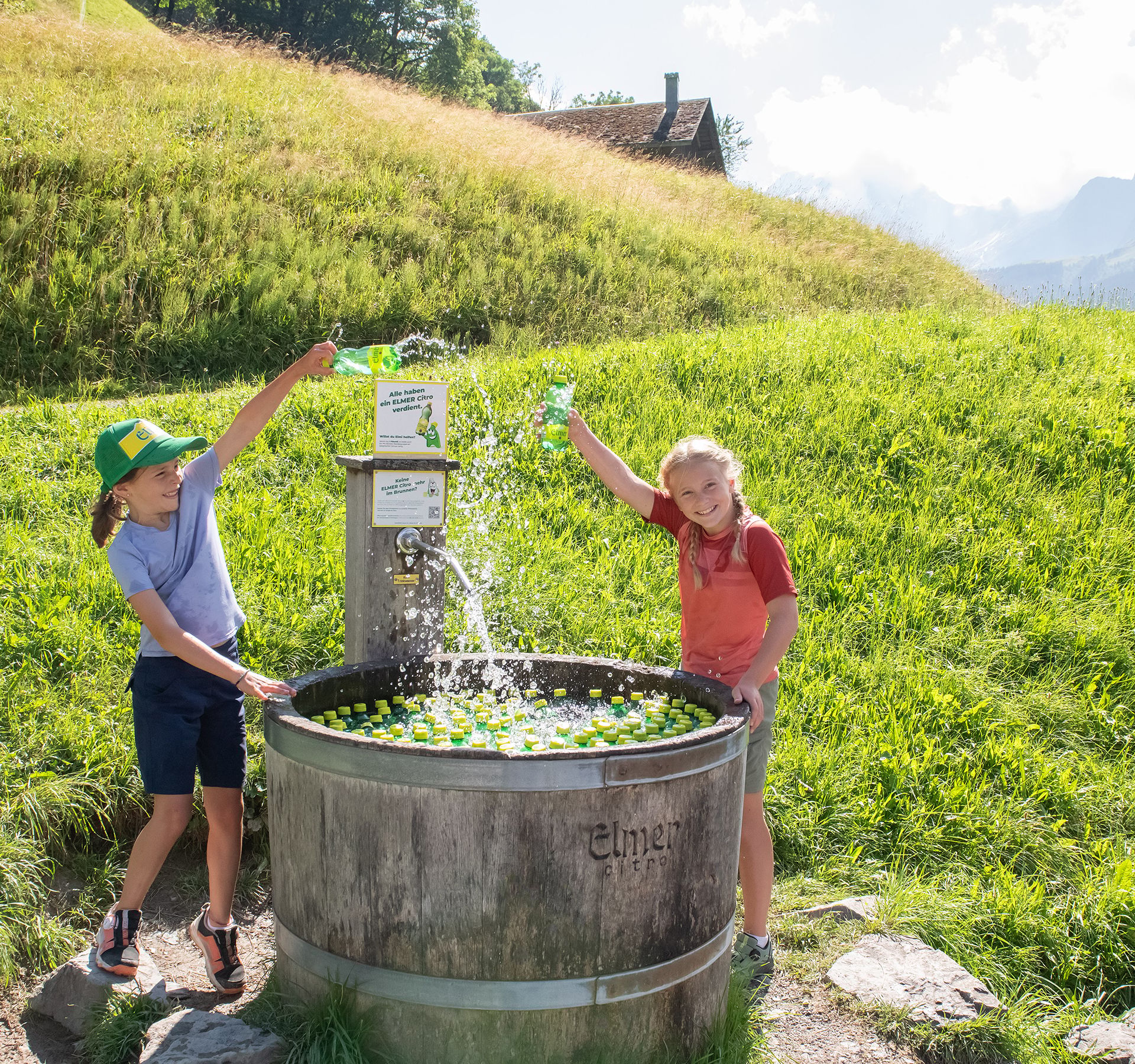 Deux jeunes filles tirent joyeusement une petite bouteille d'Elmer Citro d'une grande fontaine dans laquelle flottent de nombreuses autres bouteilles d'Elmer Citro...