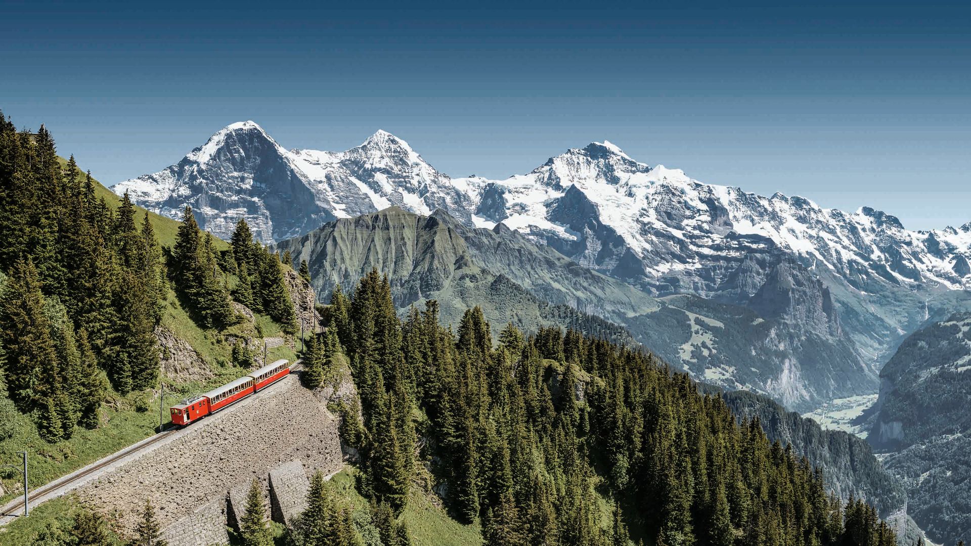 Aerial view of the cogwheel going up the mountain. In the background, snow-covered peaks of the Eiger, Mönch and Jungfrau can be seen.