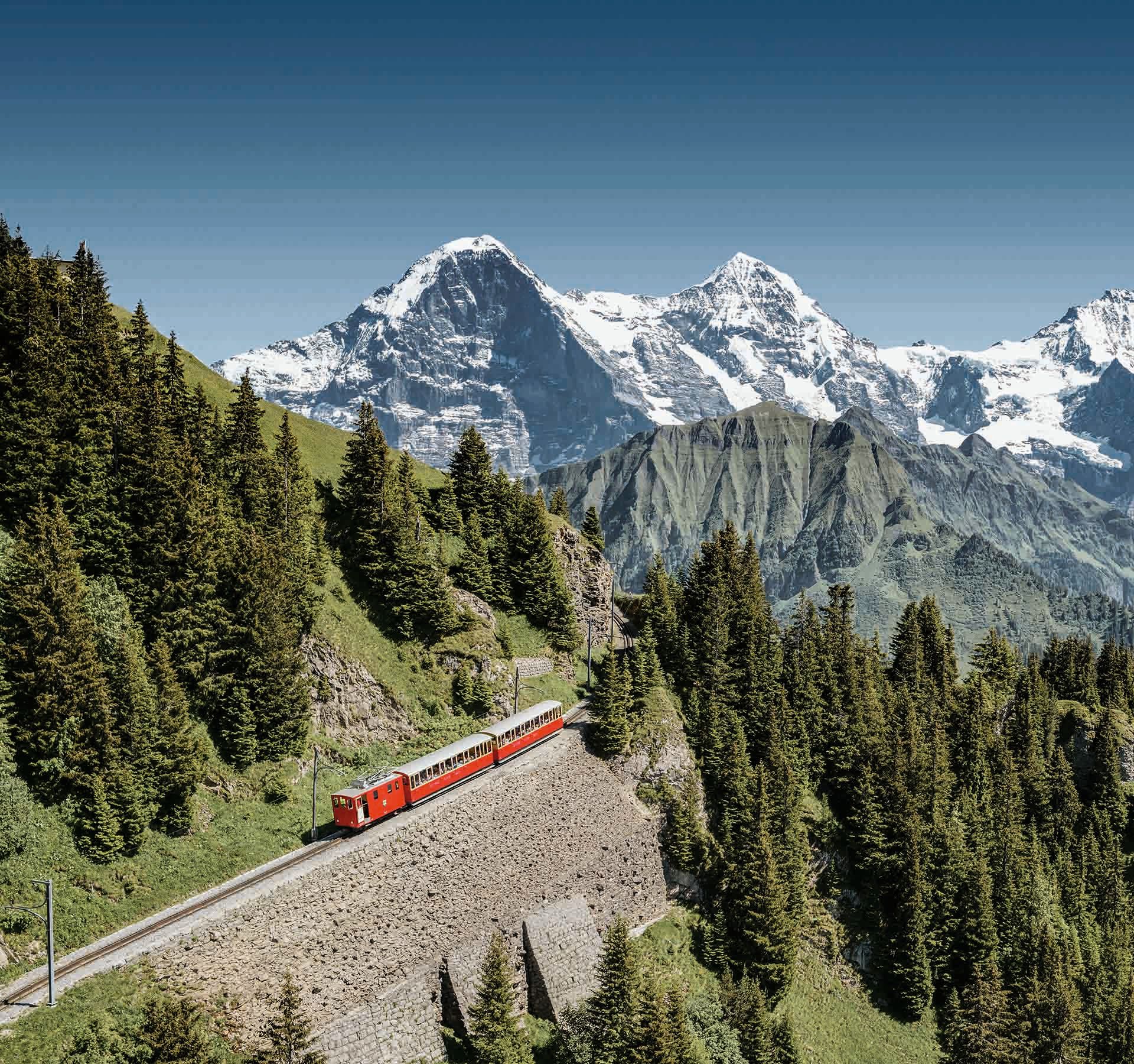 Aerial view of the cogwheel going up the mountain. In the background, snow-covered peaks of the Eiger, Mönch and Jungfrau can be seen.