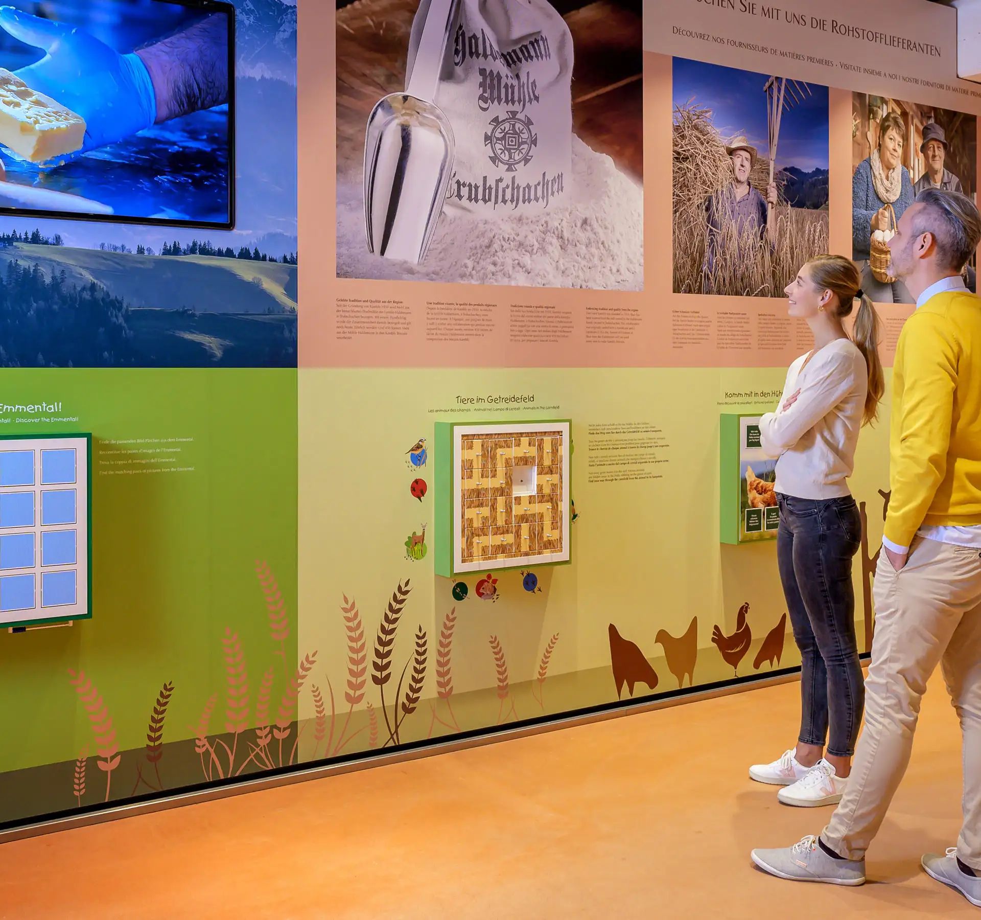 Deux personnes regardent une exposition avec des textes, des images et des éléments interactifs sur l'origine des produits Kambly dans un musée ou un centre d'accueil.