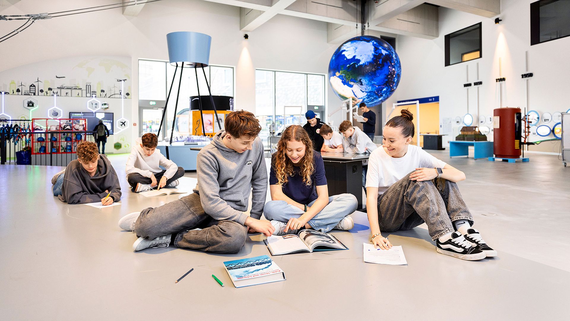A group of young people are sitting on the floor in a modern learning space, working together with books and worksheets; in the background, a large globe and interactive exhibition elements can be seen.