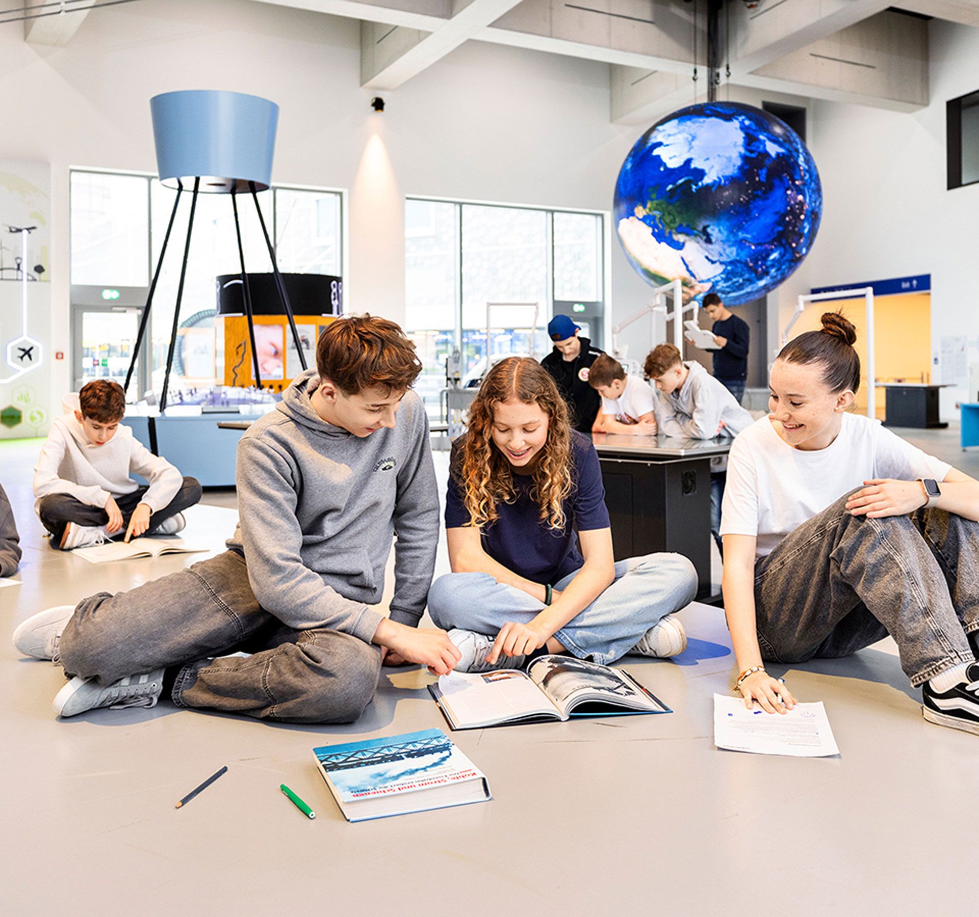 Un gruppo di giovani è seduto sul pavimento di una moderna aula didattica e lavora insieme su libri e fogli di lavoro; sullo sfondo si vedono un grande mappamondo ed elementi espositivi interattivi.