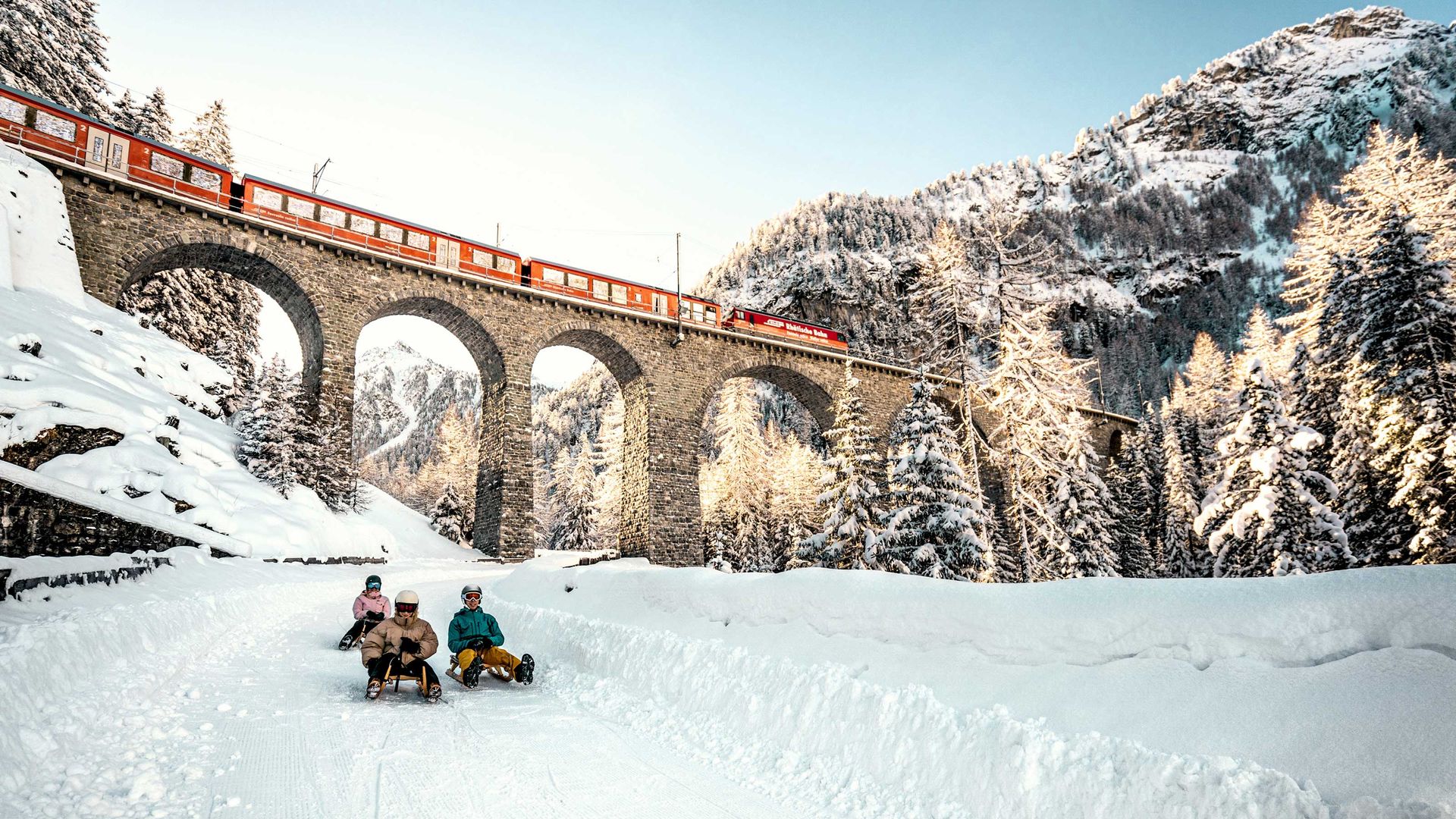 3 Personen fahren auf Schlitten die Piste hinunter. Im Hintergrund ist eine grosse Eisenbahnbrücke mit einem Zug darauf zu sehen. Es ist ein herrlicher Wintersporttag.