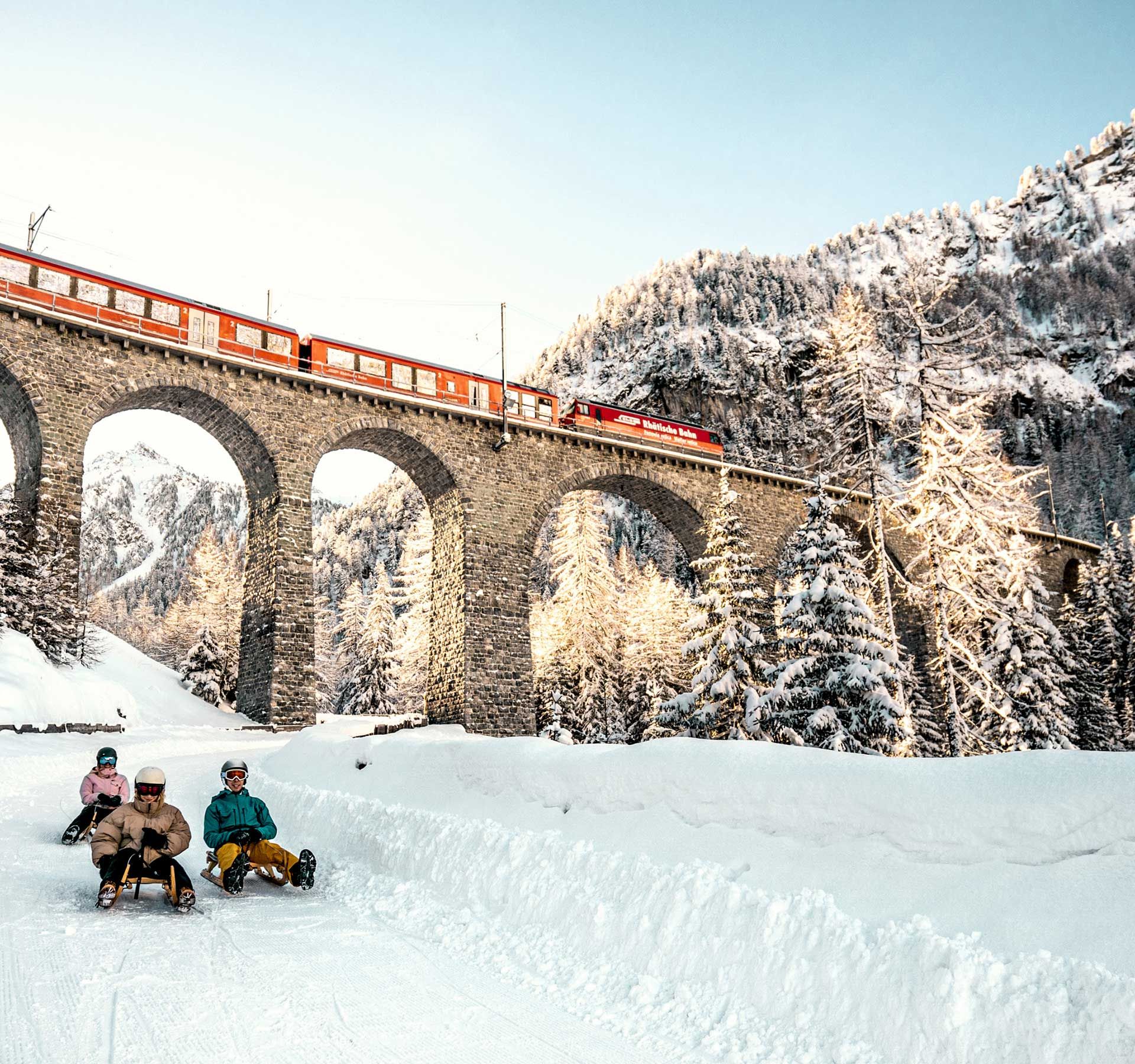 3 persone scendono dalla pista con la slitta. Sullo sfondo si vede un grande ponte ferroviario con un treno. È una meravigliosa giornata di sport invernali.