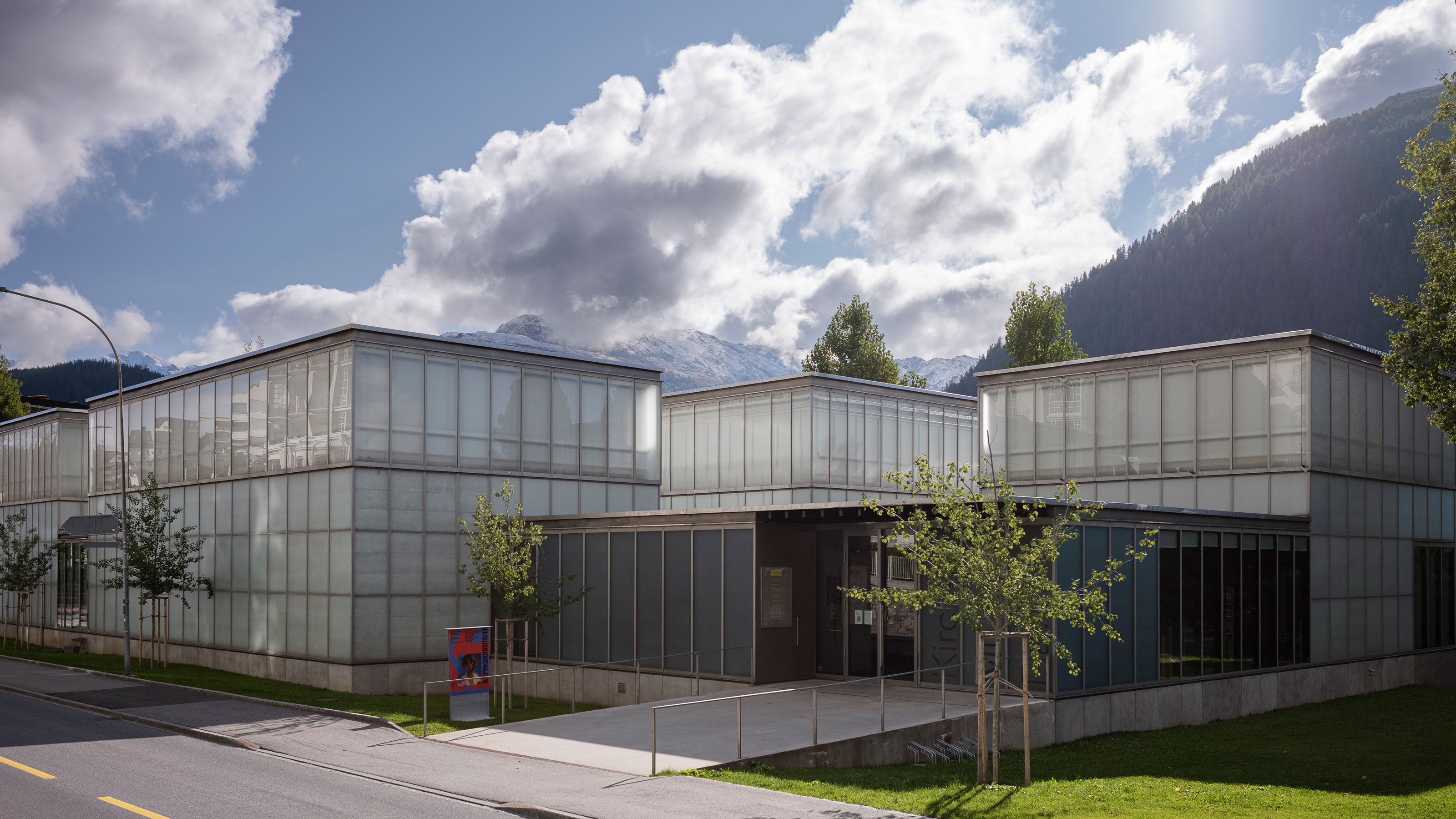 The modern building is located between trees, mountains, and a green lawn.