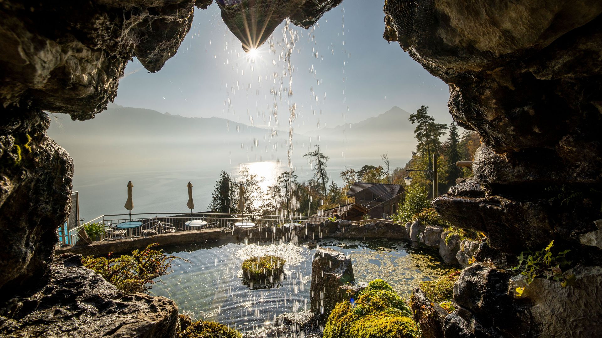 Vue depuis la grotte Saint-Beatus sur une cascade qui se jette dans un étang recouvert de mousse, avec un lac, des arbres et des montagnes en arrière-plan, sous le soleil.
