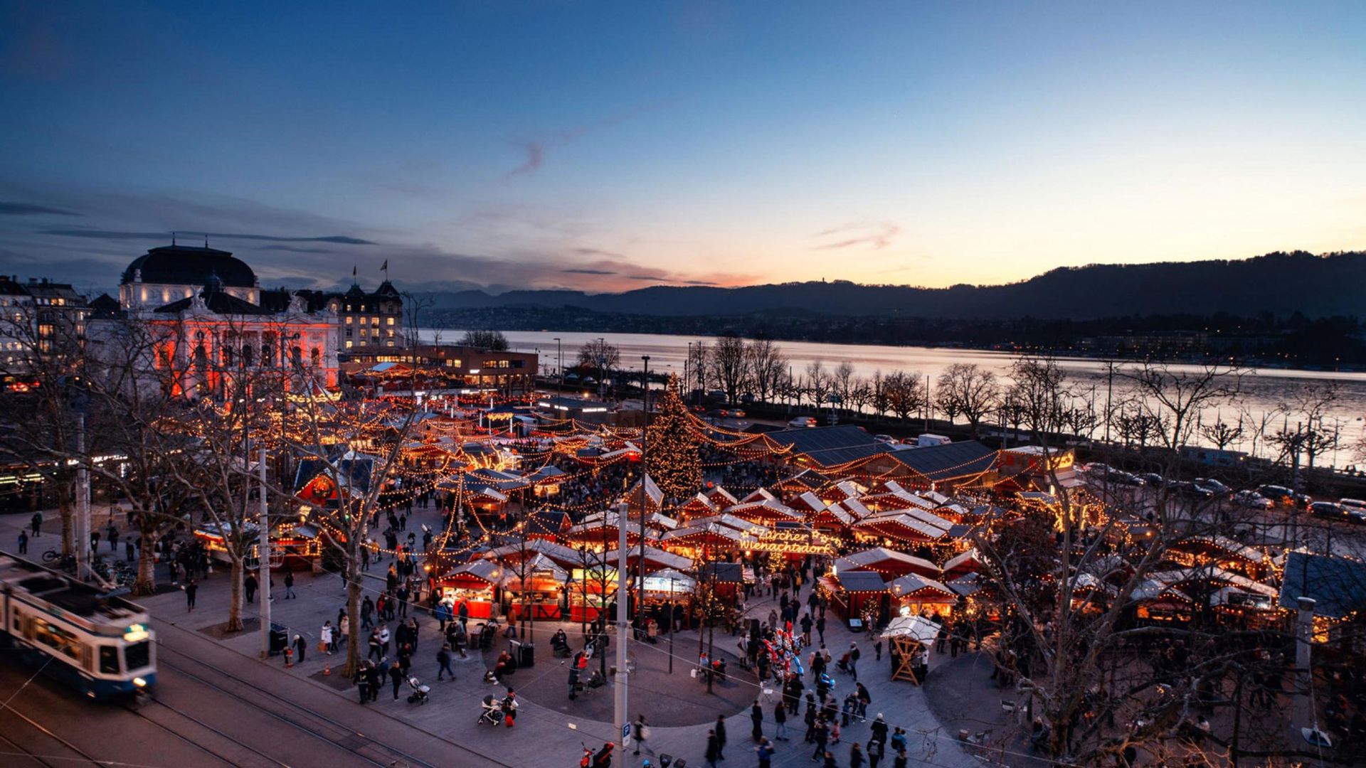 La photo montre le village de Noël de Zurich illuminé au crépuscule, avec de nombreux stands de marché, des lumières et des gens qui visitent le marché de Noël.