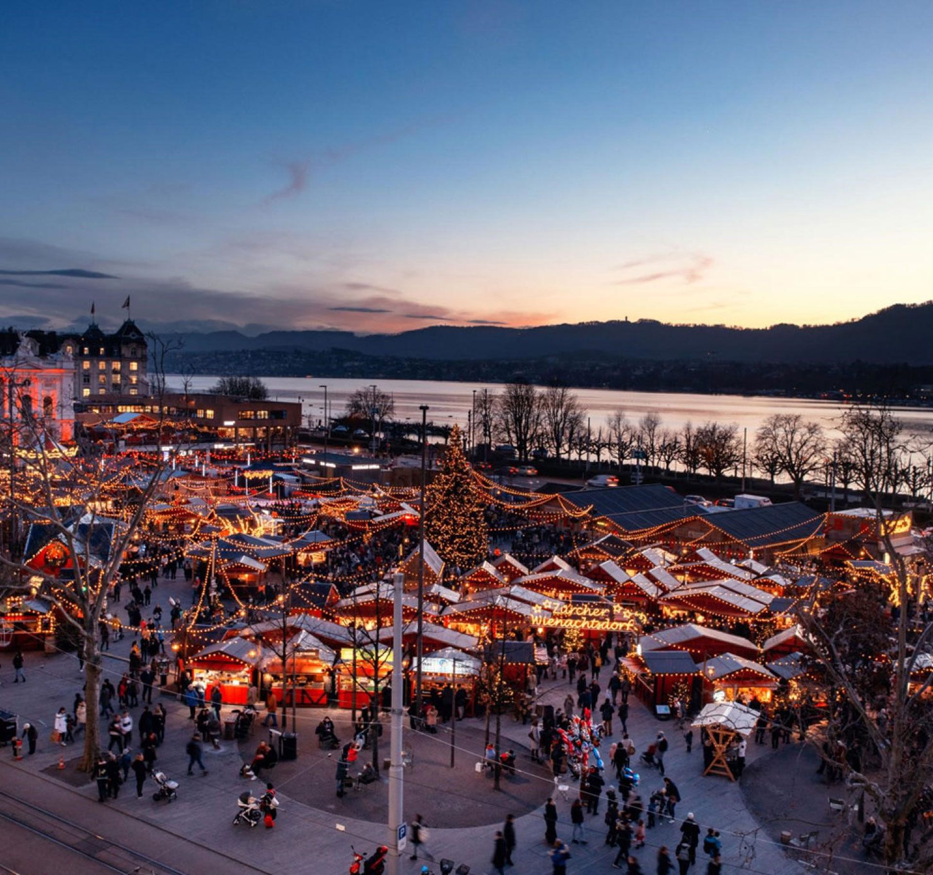 La photo montre le village de Noël de Zurich illuminé au crépuscule, avec de nombreux stands de marché, des lumières et des gens qui visitent le marché de Noël.