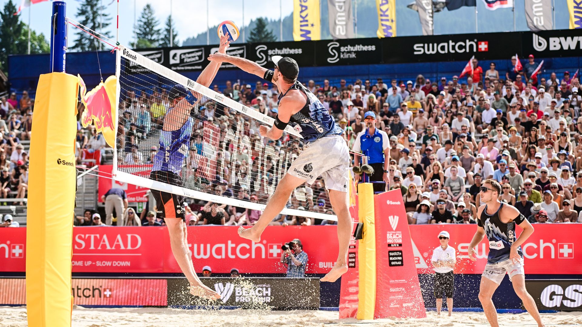Players competing at the Swatch Beach Pro in Gstaad on the sand court, with spectator stands and mountains in the background, Swiss Beach Volleyball event.