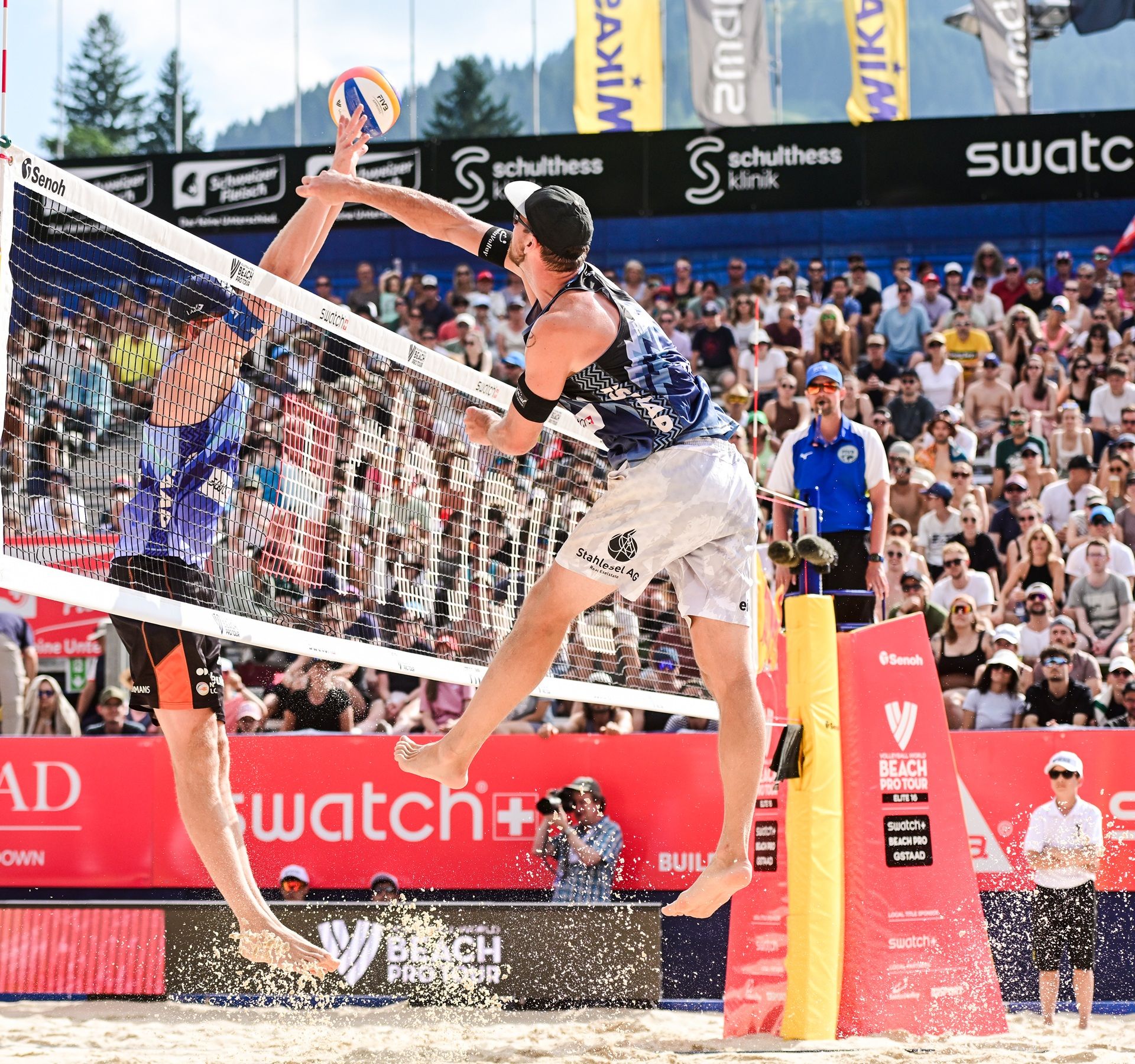 Players competing at the Swatch Beach Pro in Gstaad on the sand court, with spectator stands and mountains in the background, Swiss Beach Volleyball event.