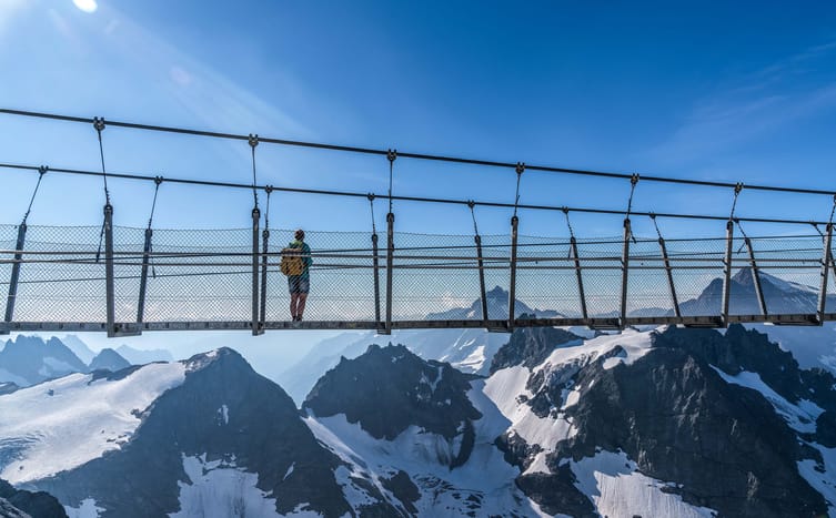 Eine Person steht auf einer Hängebrücke und blickt auf eine majestätische Berglandschaft.