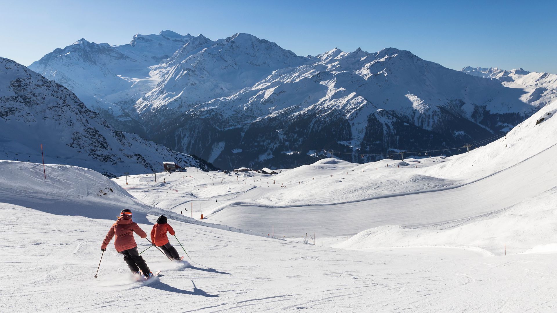 Zwei Skifahrer fahren auf einer präparierten Piste im Skigebiet 4 Vallés, umgeben von schneebedeckten Bergen unter klarem Himmel.