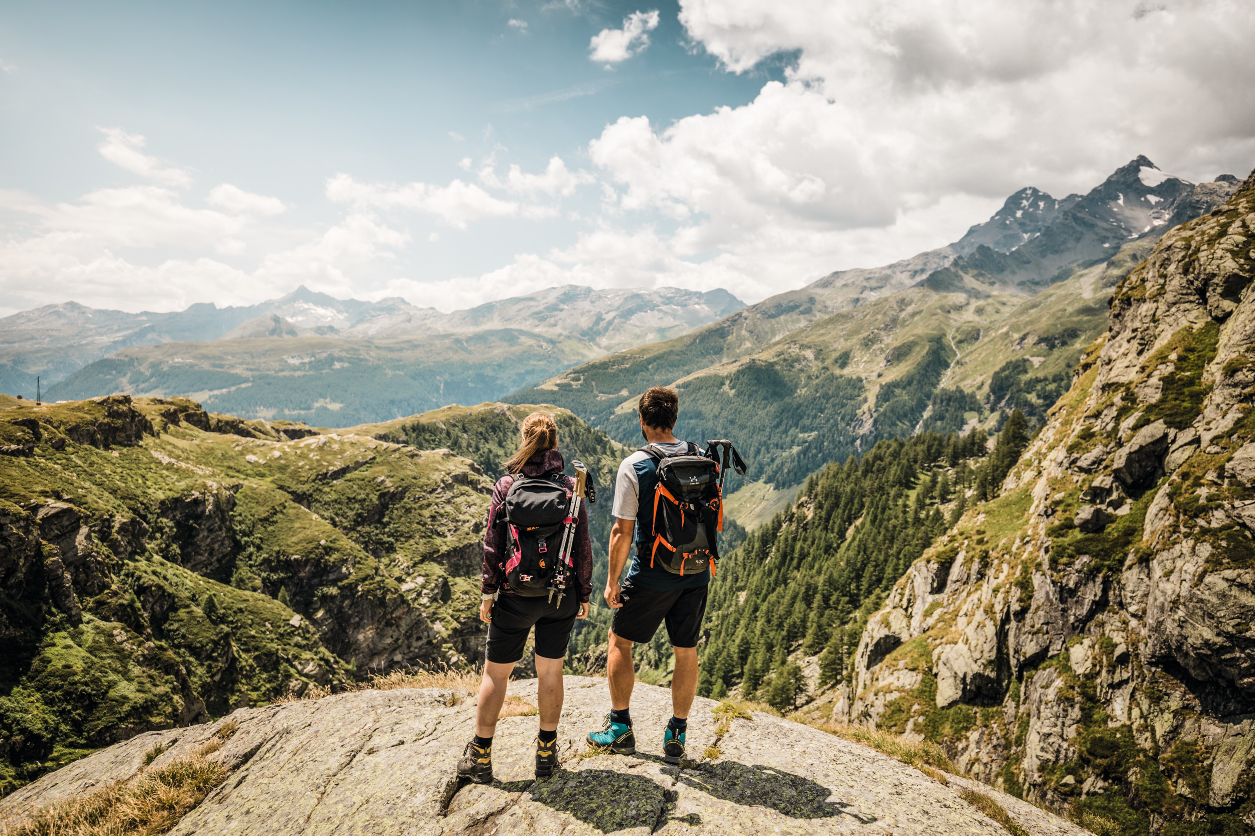 Un couple profite d'une vue magnifique sur les montagnes. Tous deux portent un sac à dos et sont équipés pour la randonnée. 