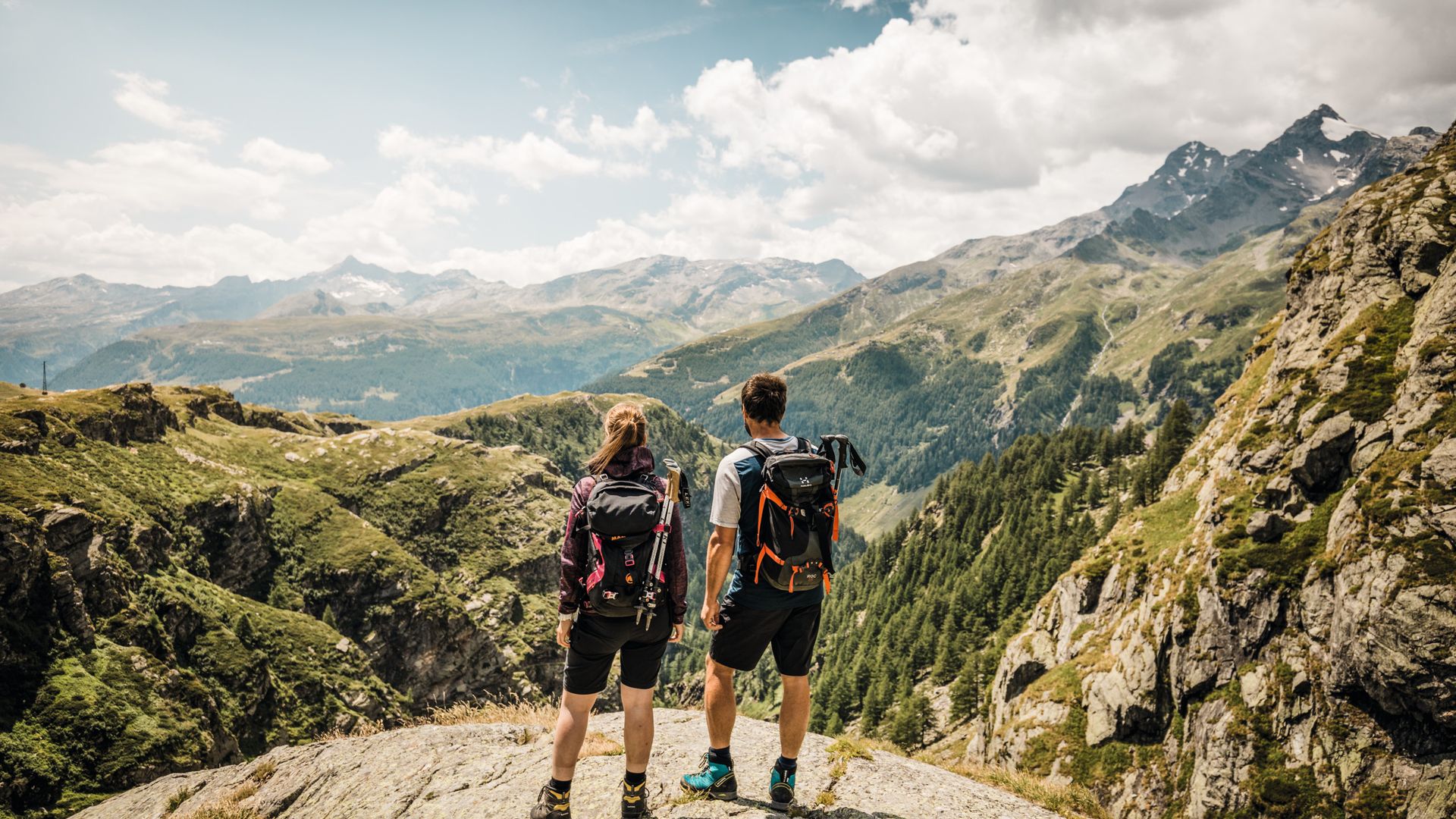 A couple enjoys the wonderful view of the mountains. Both are carrying a backpack and hiking gear. 